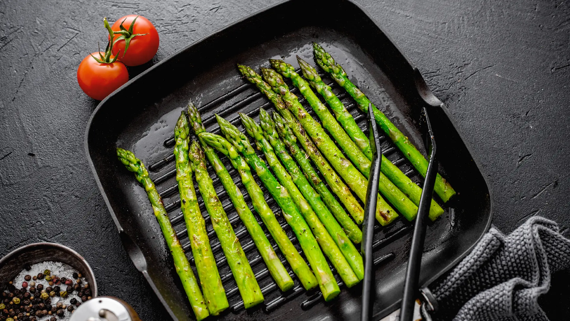 A top-down shot of a black grill pan filled with green asparagus spears being cooked. Two tomatoes sit next to the pan.