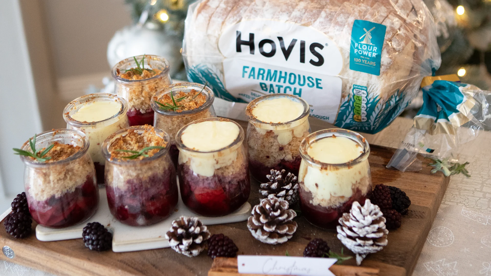 A close-up of hot custard being poured into a jar of golden baked fruit crumble, with a loaf of Hovis® Farmhouse Batch in the background - the bread used to make the buttery crumble topping.