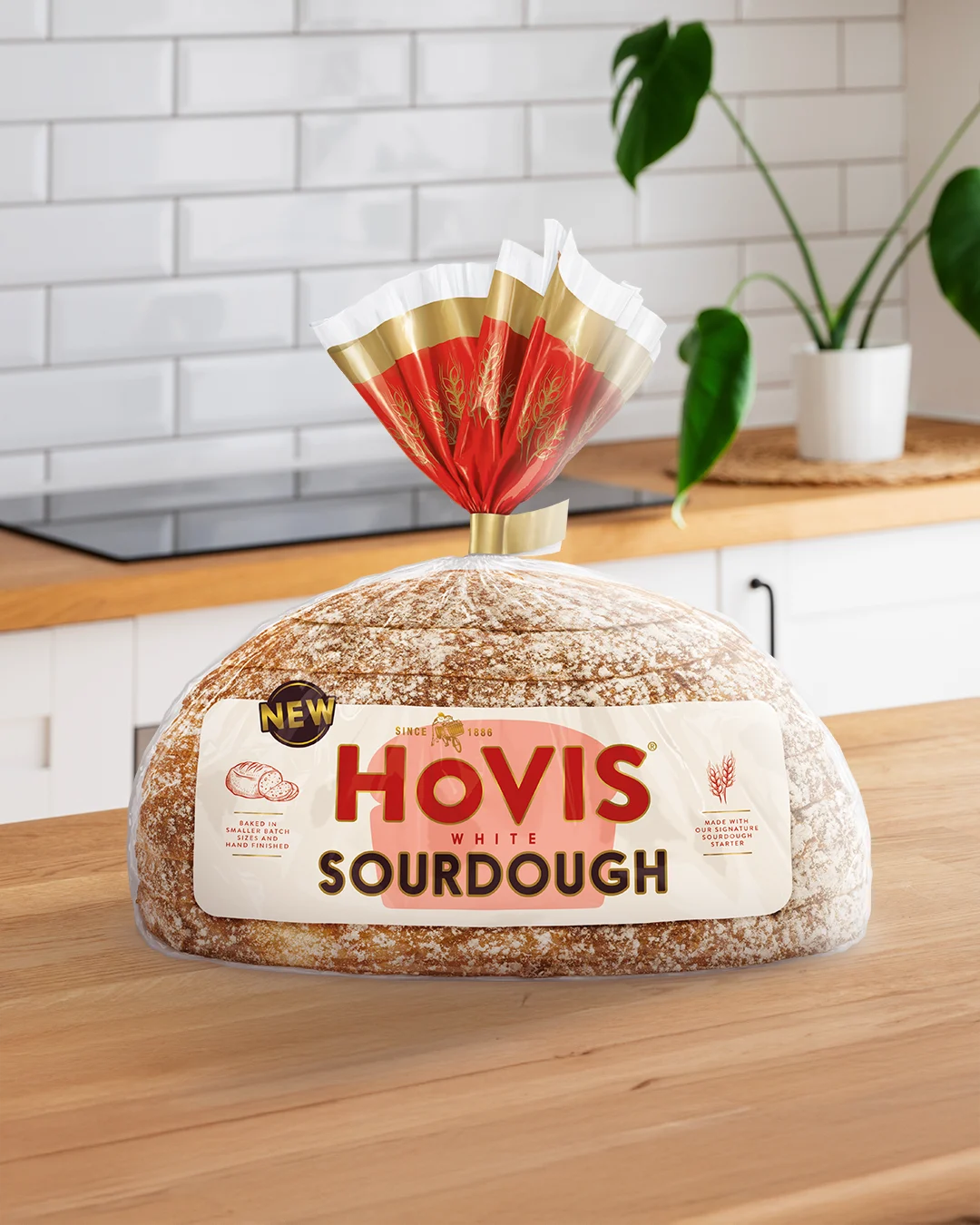 A loaf of Hovis White Sourdough bread displayed on a wooden kitchen countertop. The packaging features a white and red design with a gold trim and nutrition information. A 'NEW' label is shown on the top corner of the pack