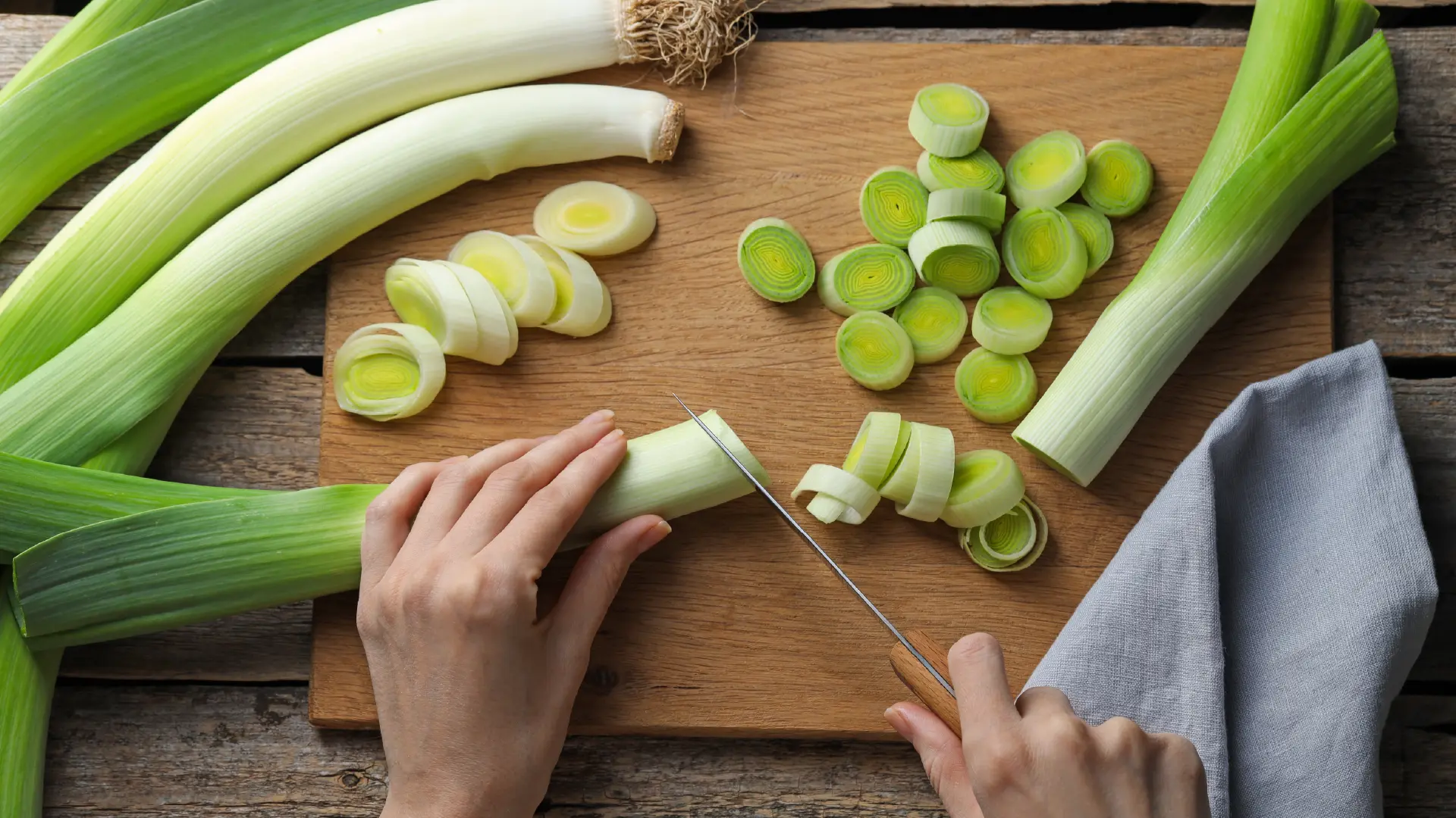 Hands slicing fresh leeks into rounds on a wooden cutting board, surrounded by whole leeks and a grey kitchen towel.