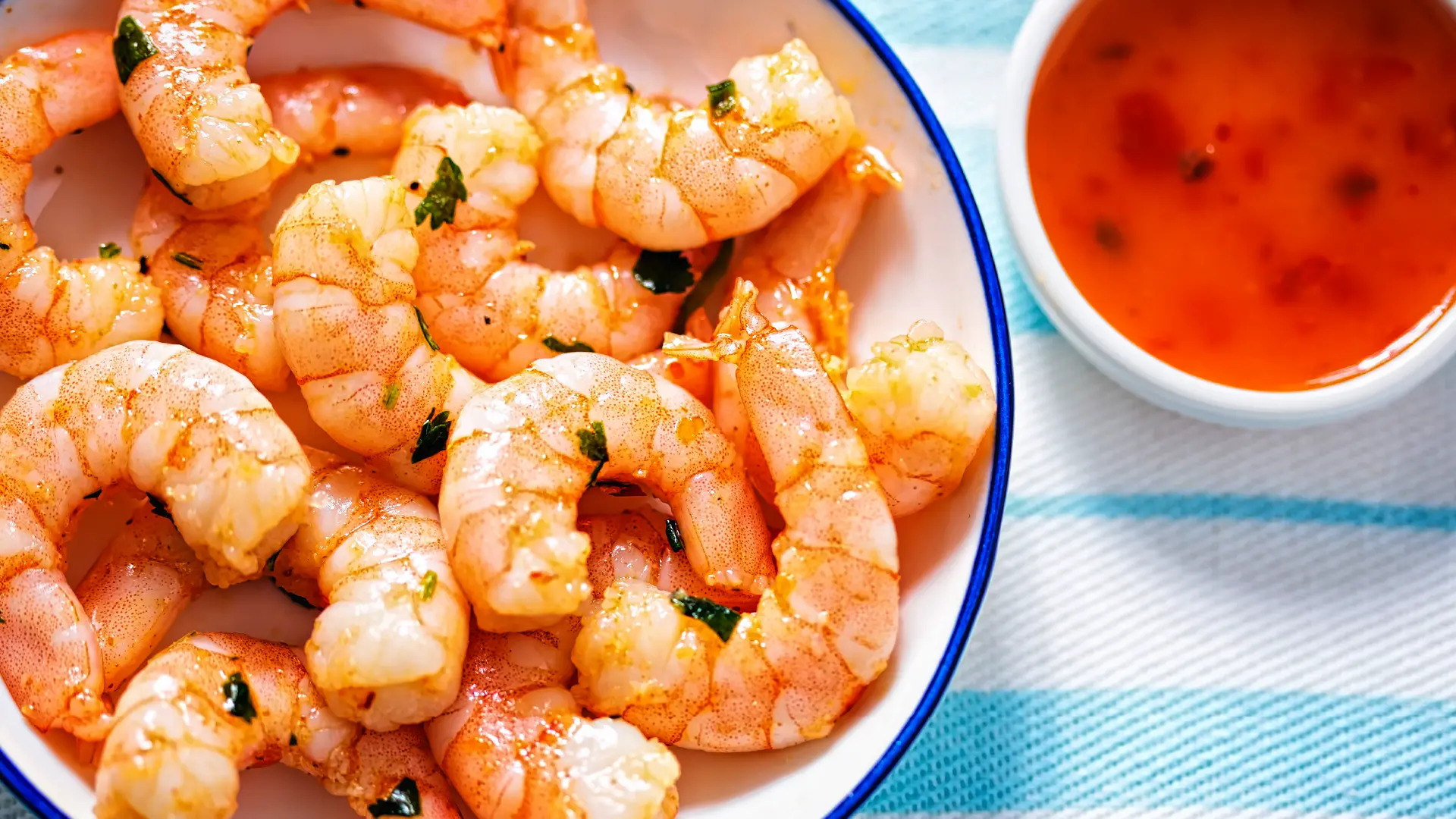A top-down view of a plate filled with raw prawns topped with chopped greens and seasoning, with a small bowl of chilli sauce on the side.