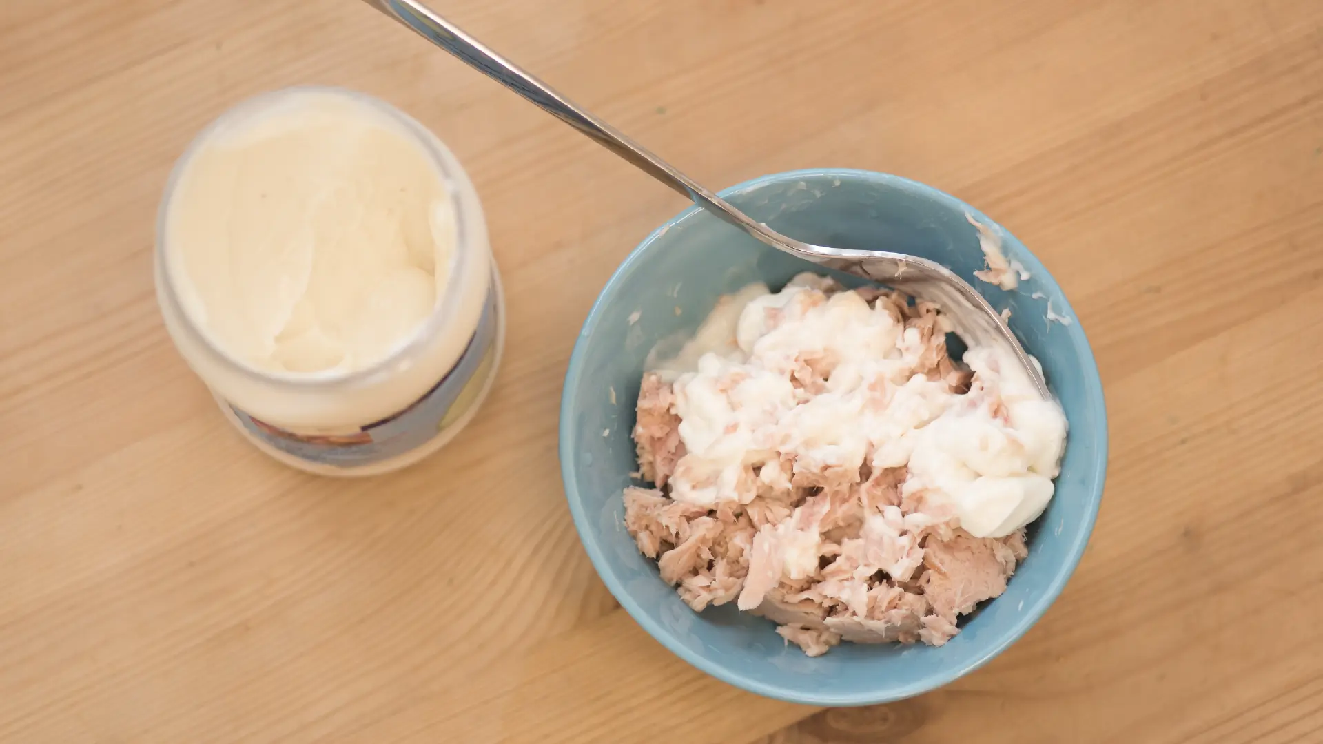 Overhead view of a light blue bowl with flaked tuna and mayonnaise, a fork inside, next to an open jar of mayonnaise.