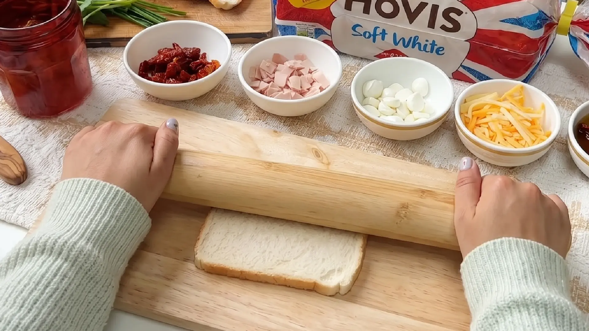 A person uses a wooden rolling pin to flatten a slice of white bread, surrounded by small bowls of toppings.
