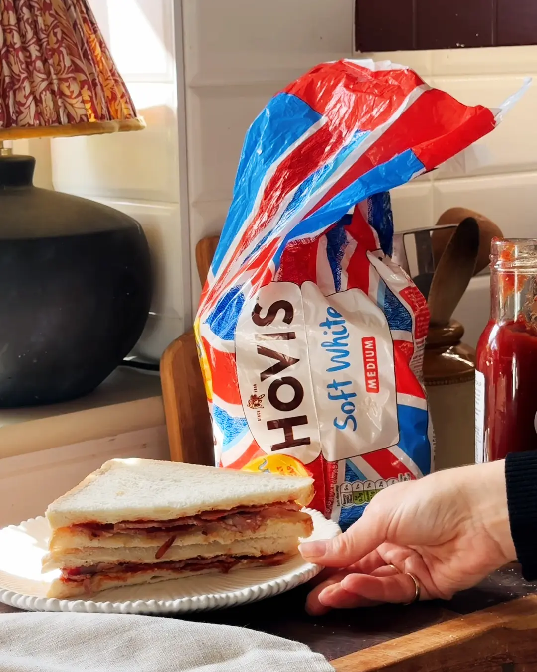 A hand next to a double-decker bacon sandwich on a plate, with a loaf of Hovis Soft White bread standing in the background.
