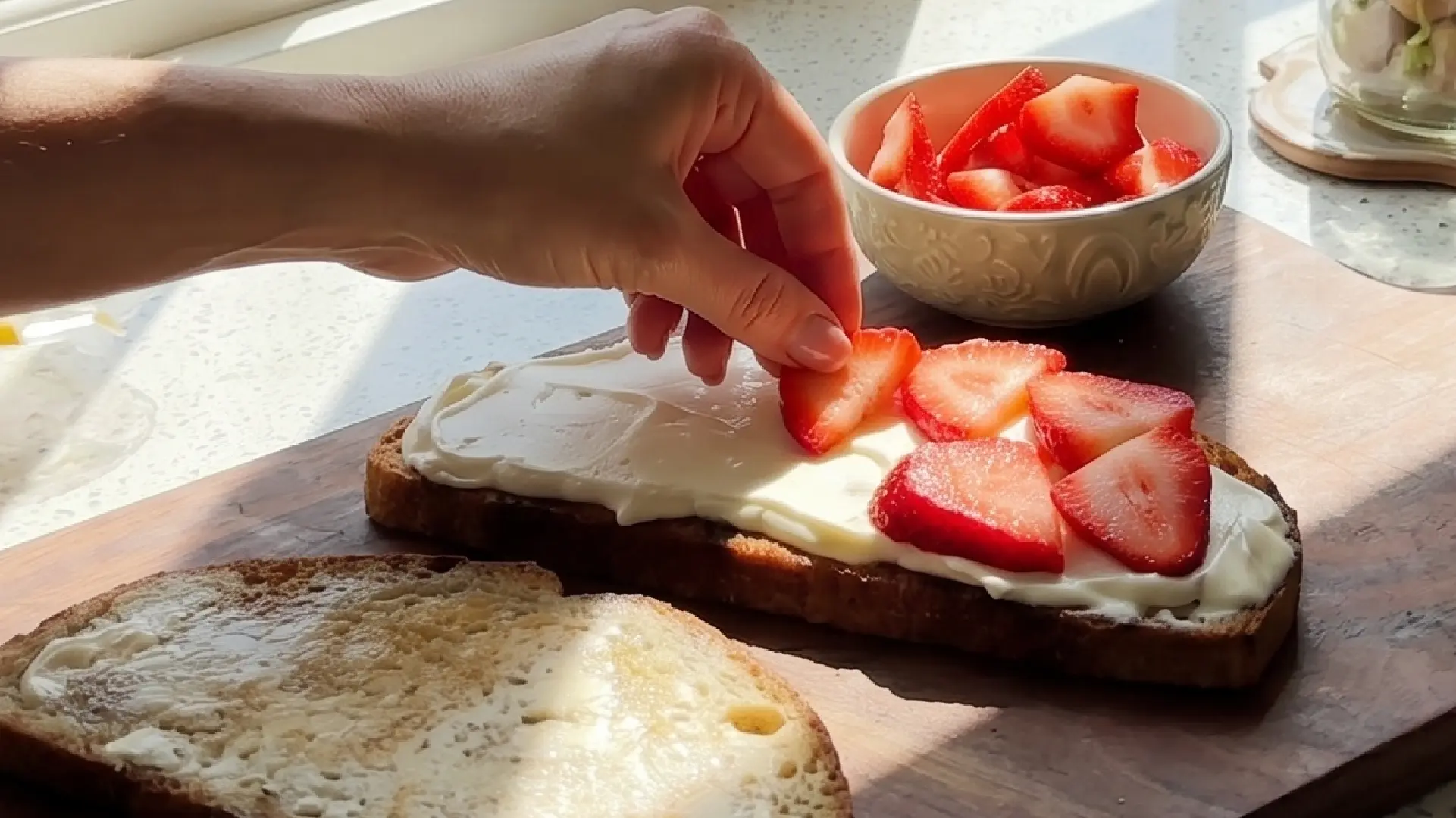 A hand places fresh, sliced strawberries onto a piece of sourdough bread topped with white cream on a wooden board.