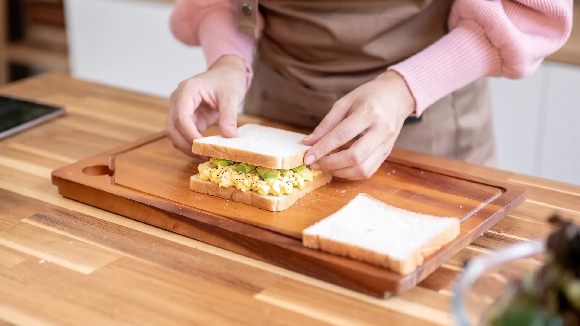 Hands placing top slice of white bread onto egg and avocado filling on a wooden cutting board.