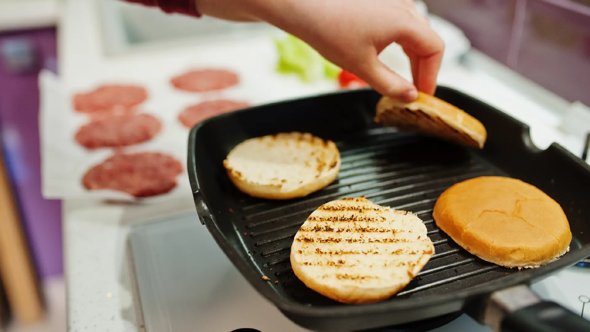 A hand flips burger buns toasting on a black grill pan, with raw burger patties visible on a sheet in the background.