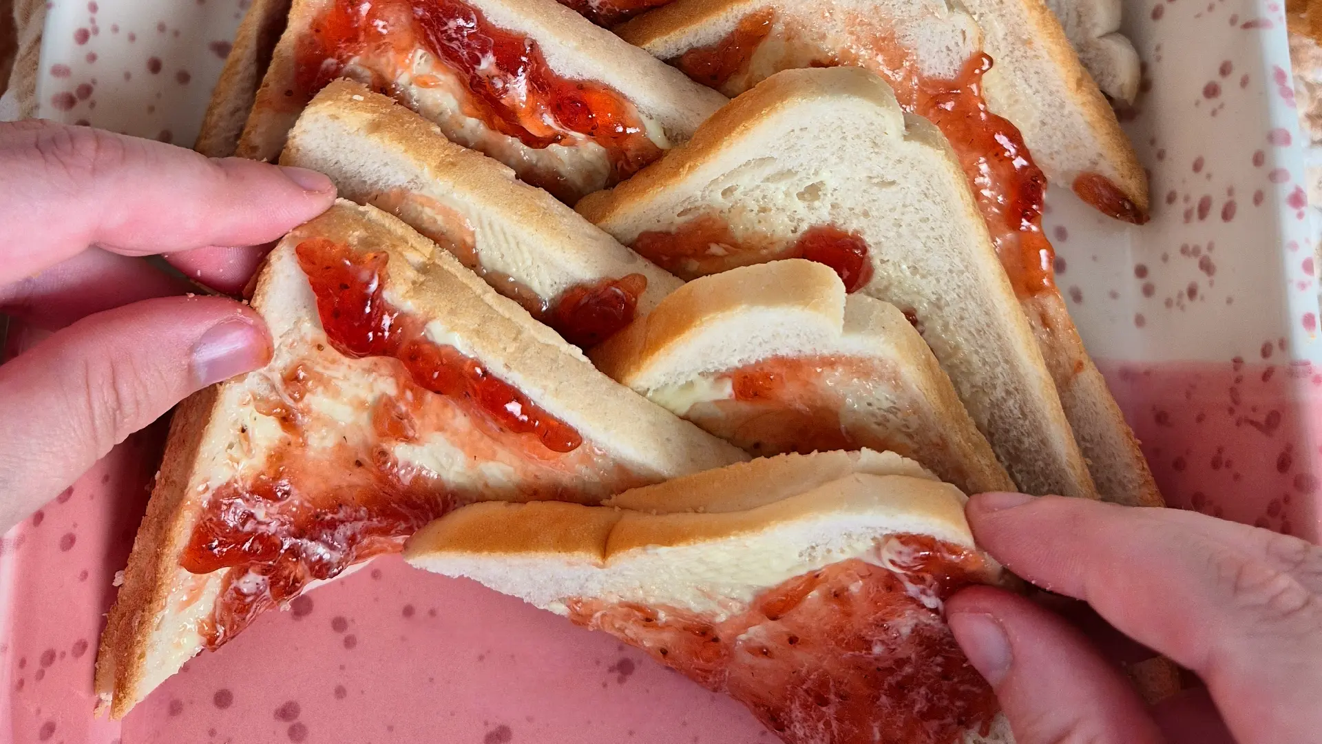 Two hands arrange triangular slices of buttered white bread spread with red jam on a speckled pink baking dish.