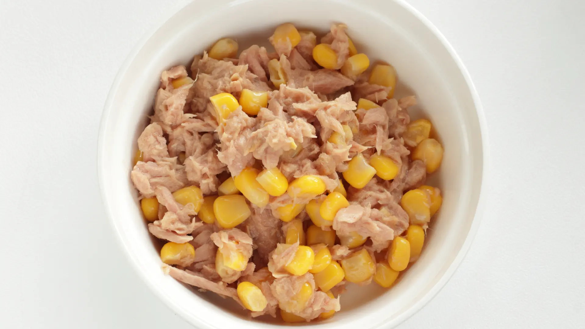 Top down view of flaked tuna mixed with bright yellow sweetcorn kernels in a white ceramic bowl.