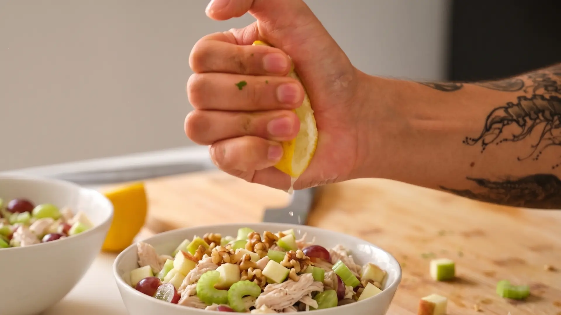 A tattooed hand squeezing fresh lemon juice over a bowl of chicken salad with walnuts, celery, apples and grapes.