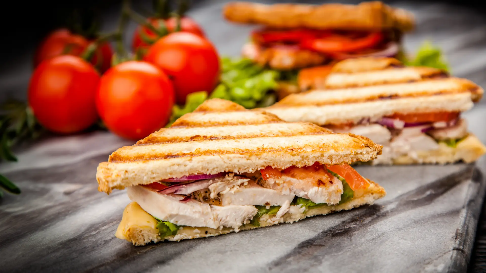 A toasted Hovis Soft White Medium Sliced sandwich, filled with tender chicken, juicy tomato slices, and fresh salad leaves, cut in half. In the background, vine tomatoes and extra sandwich slices are in the background.