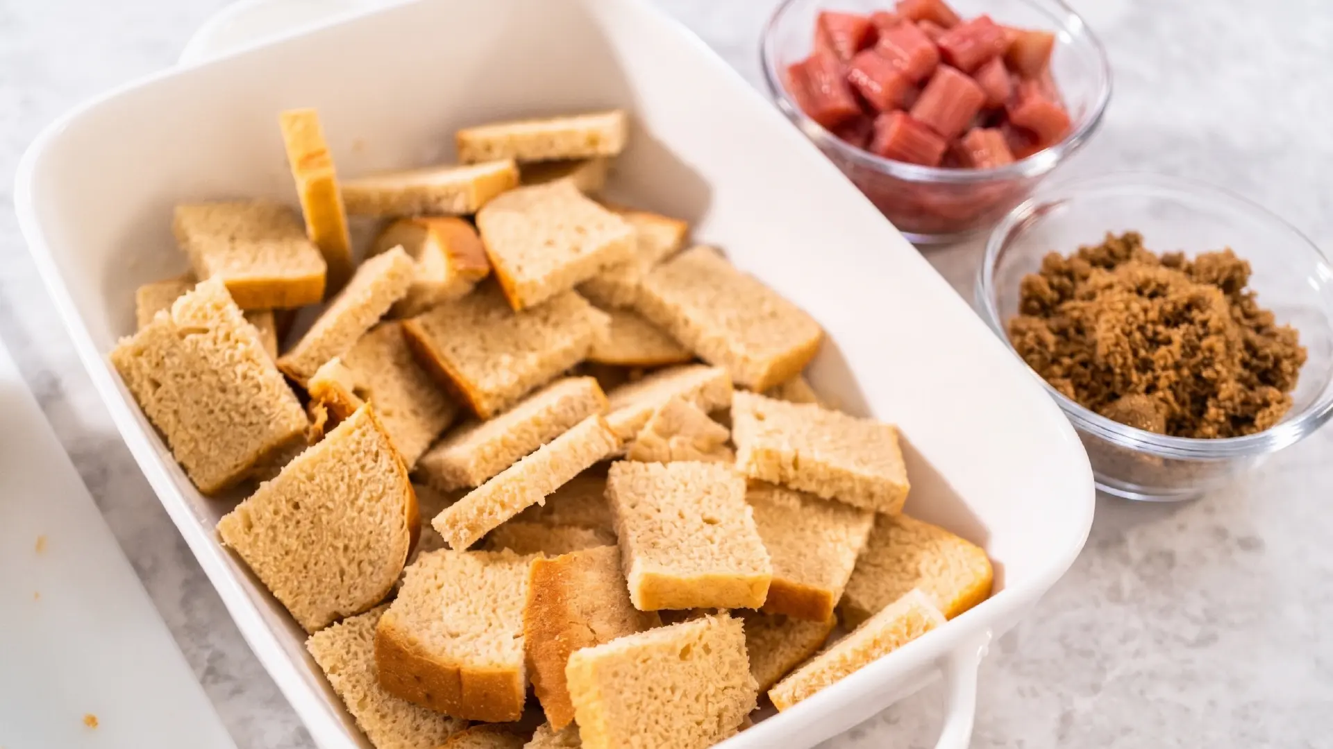 Diced bread cubes fill a white baking dish, with small bowls of chopped rhubarb and brown sugar sitting nearby.