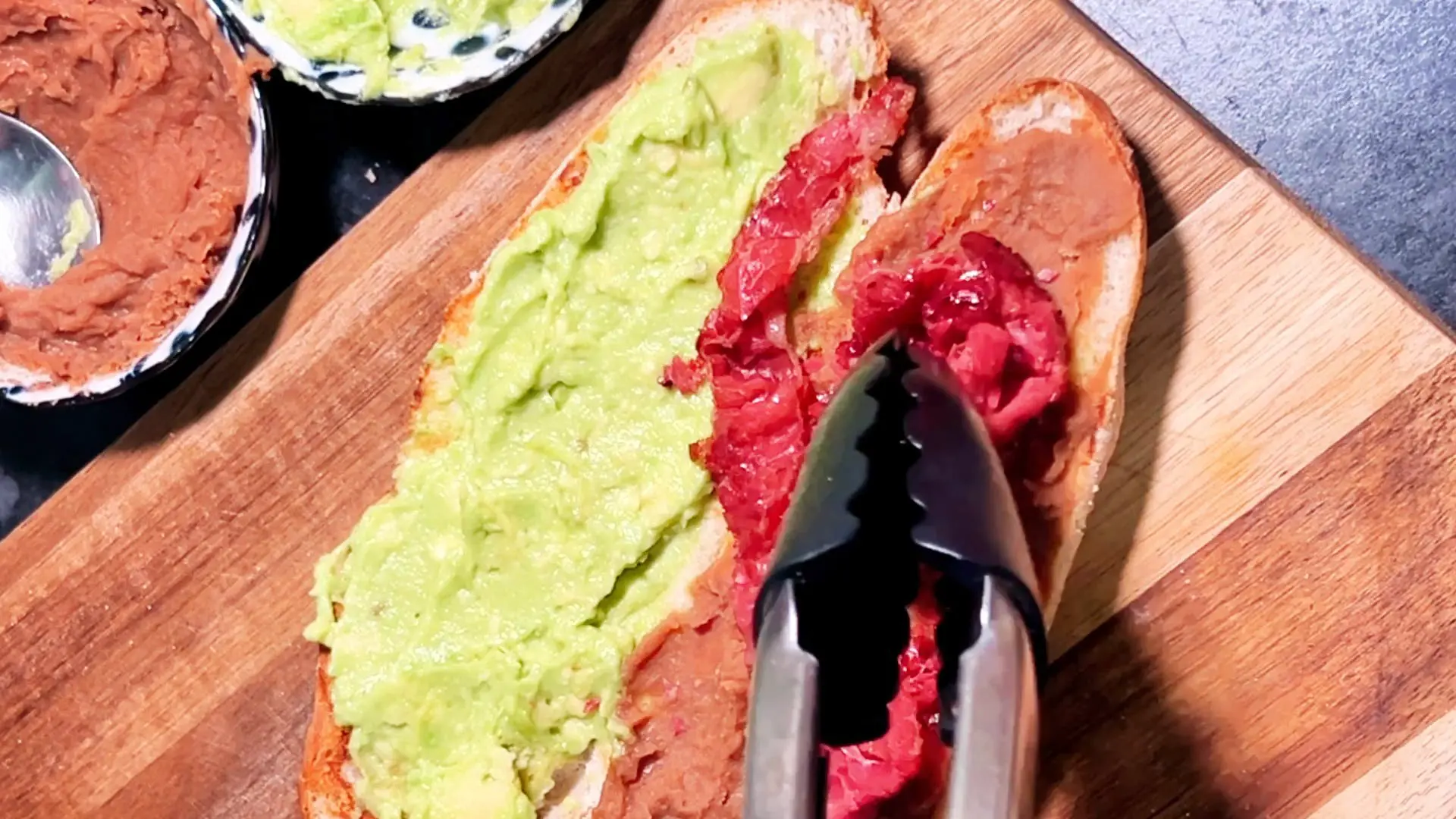 Tongs place bright red ingredients onto a baguette spread with avocado and paste, sitting on a wooden cutting board.