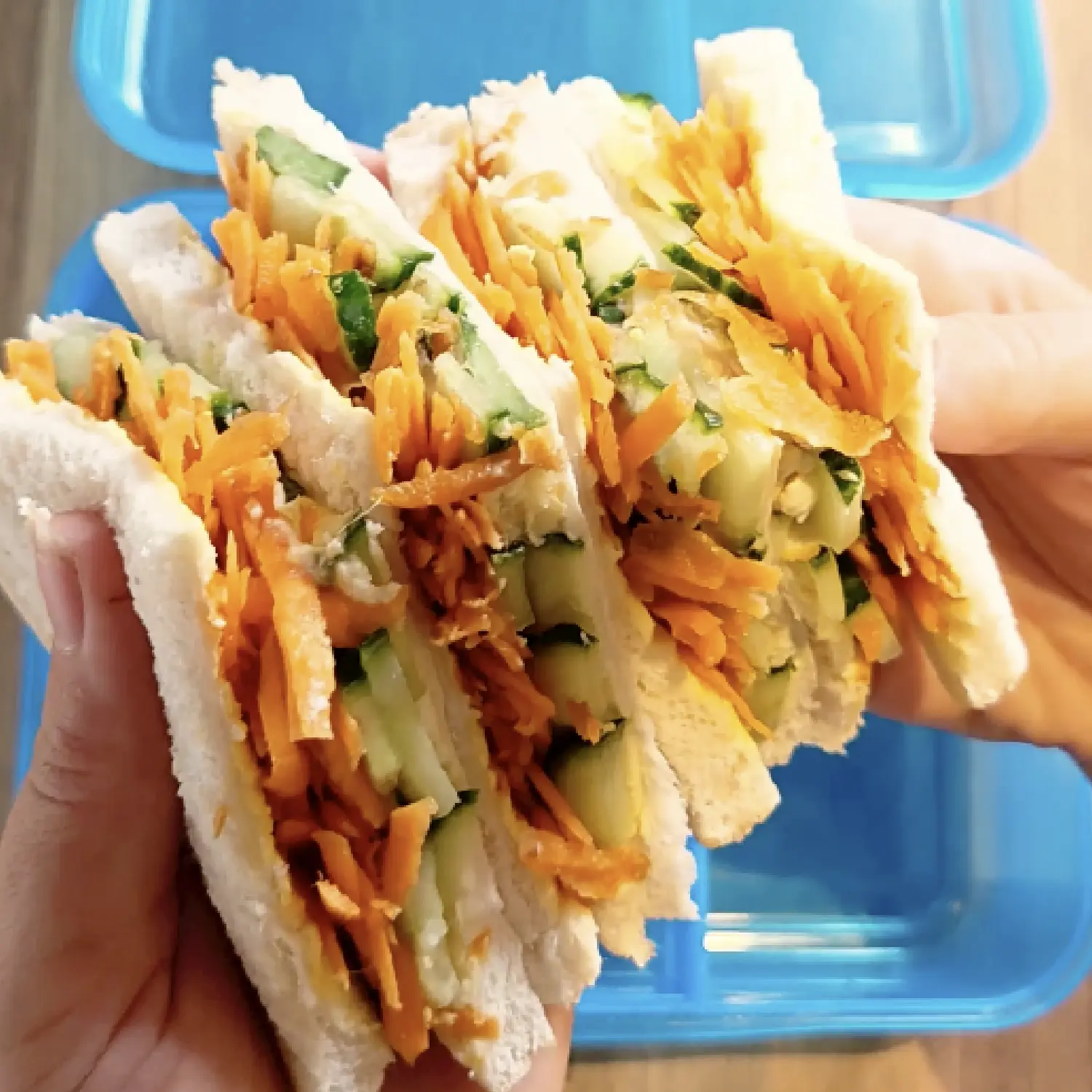 Hands holding four white bread sandwich halves stuffed with shredded carrots, cucumber slices and hummus over a blue container.