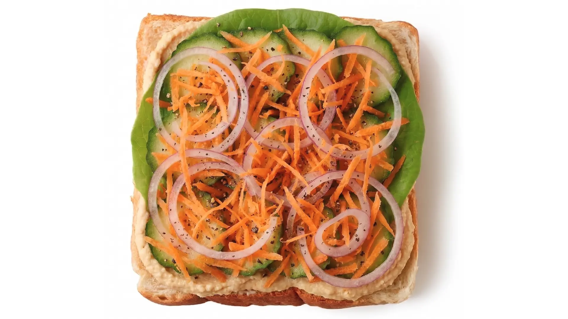Top down view of white bread topped with hummus, lettuce, cucumber, red onion and bright shredded carrots.