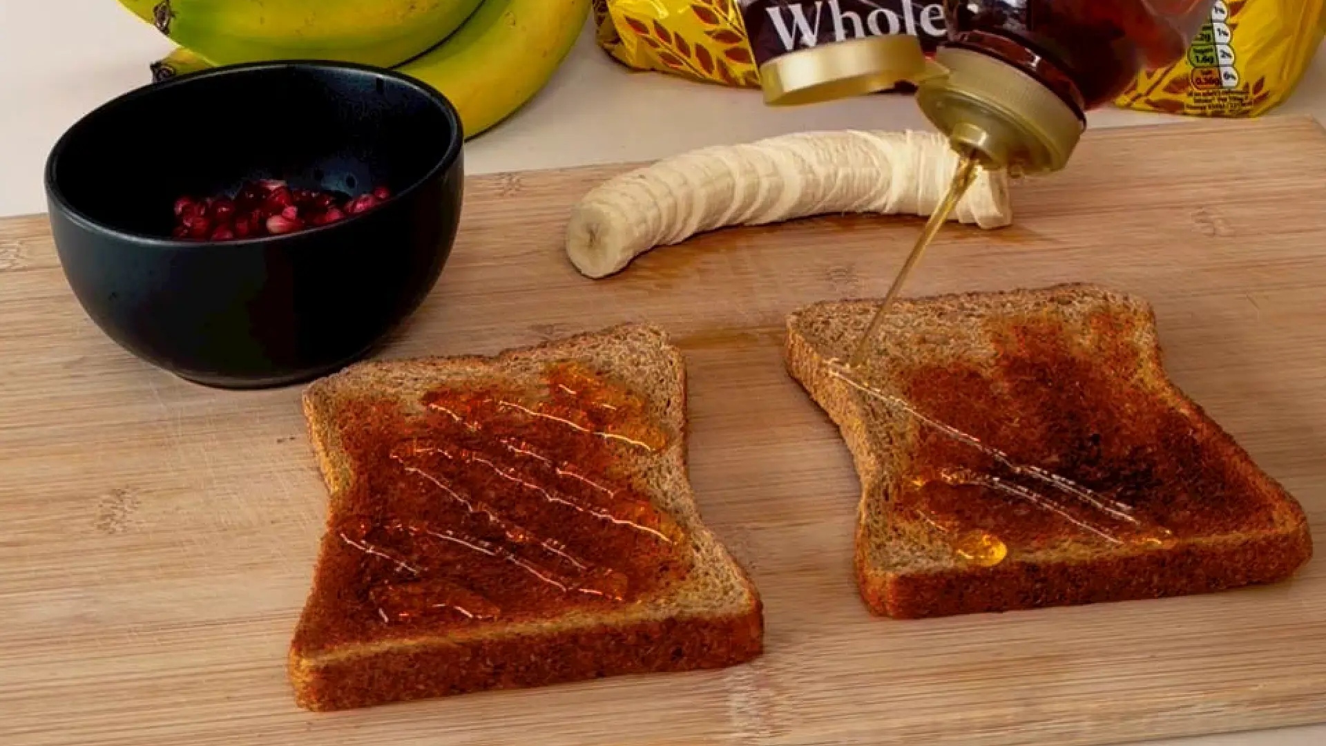 Honey drizzled over two slices of wholemeal toast on a board, with a sliced banana and pomegranate seeds in the background.