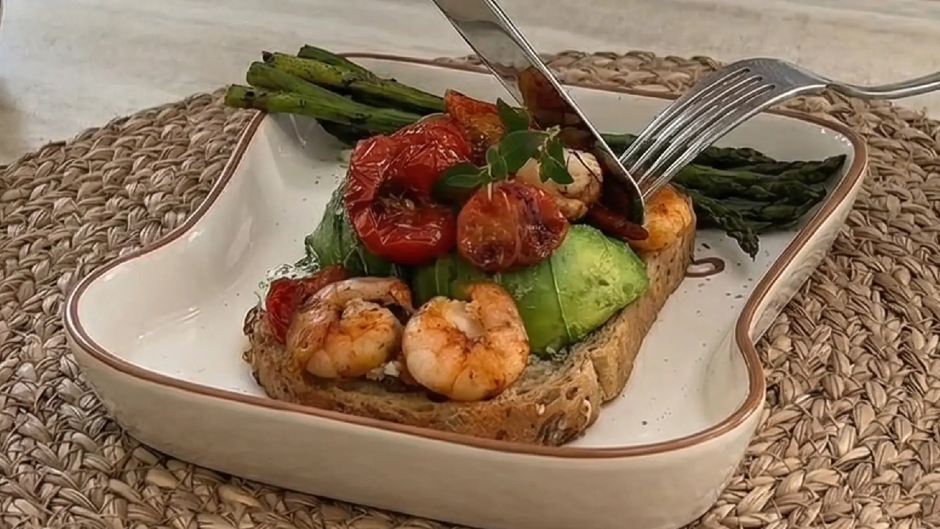 Cutting into a slice of seeded toast topped with avocado, prawns, and tomatoes, served with grilled asparagus.