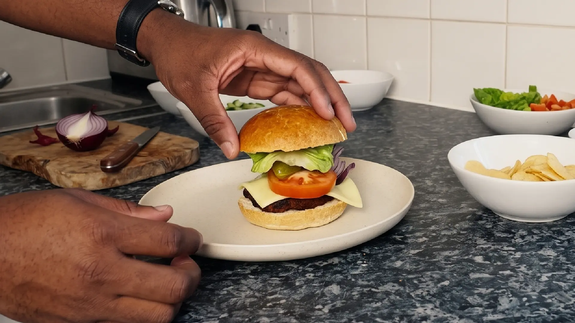 A person places the top bun on a fully assembled burger with cheese, tomato, and lettuce on a plate in a kitchen.