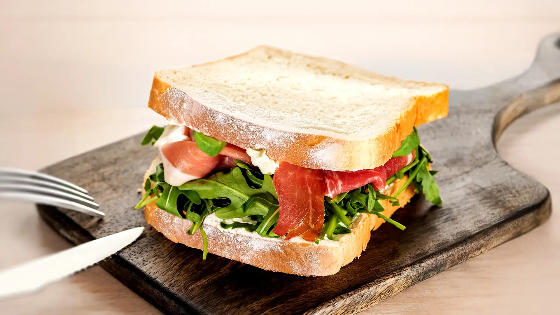 A sandwich made with Hovis Signature White bread sits on a dark wooden cutting board. The sandwich is filled with cheese spread, fresh rocket, and delicate slices of cured ham. The top slice of bread is slightly askew, revealing the delicious layers inside. A small portion of a knife and fork are visible to the left. 