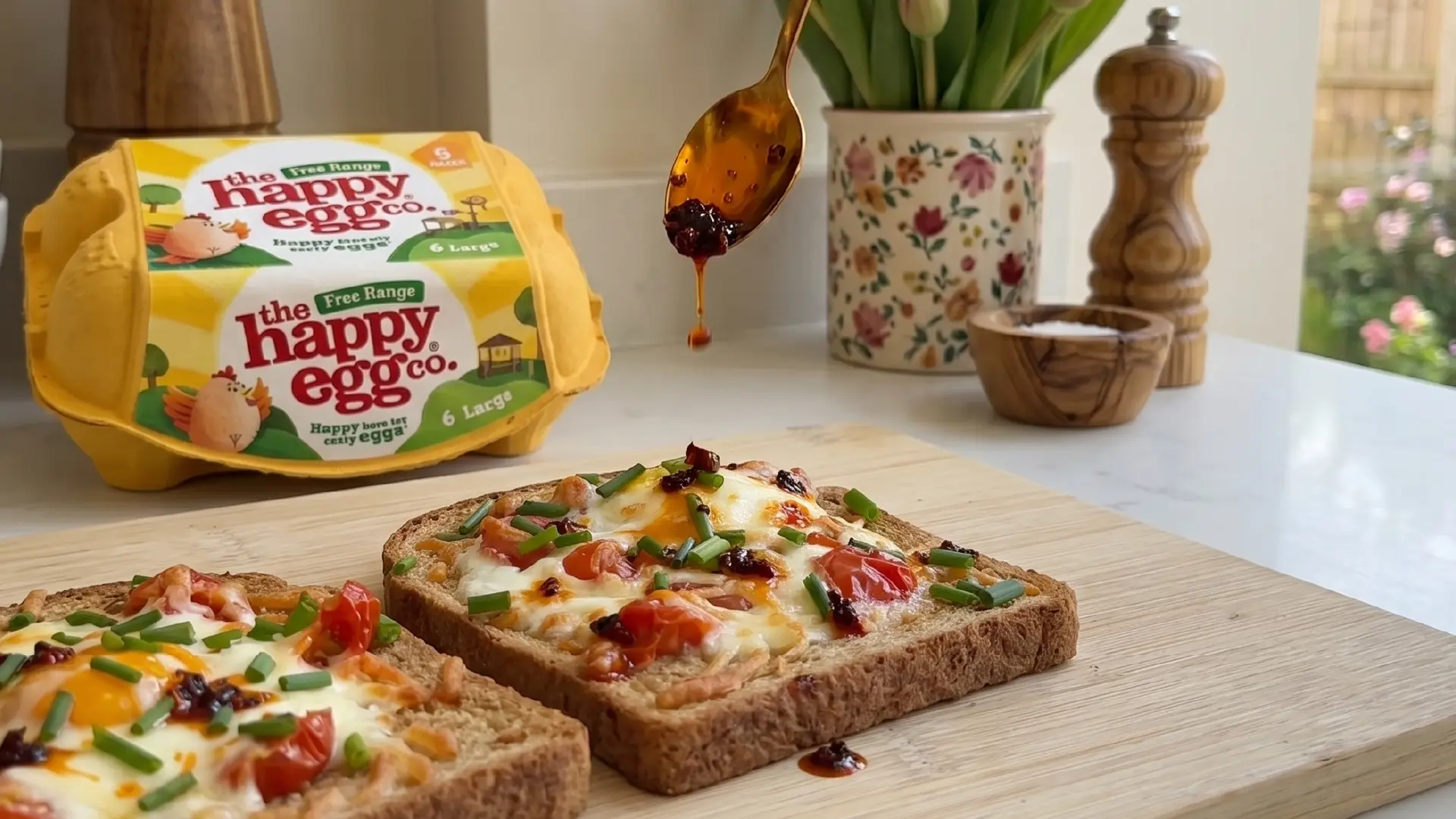 Drizzling chilli oil from a spoon onto cooked egg-in-a-hole toast with melted cheese, tomatoes and chives.