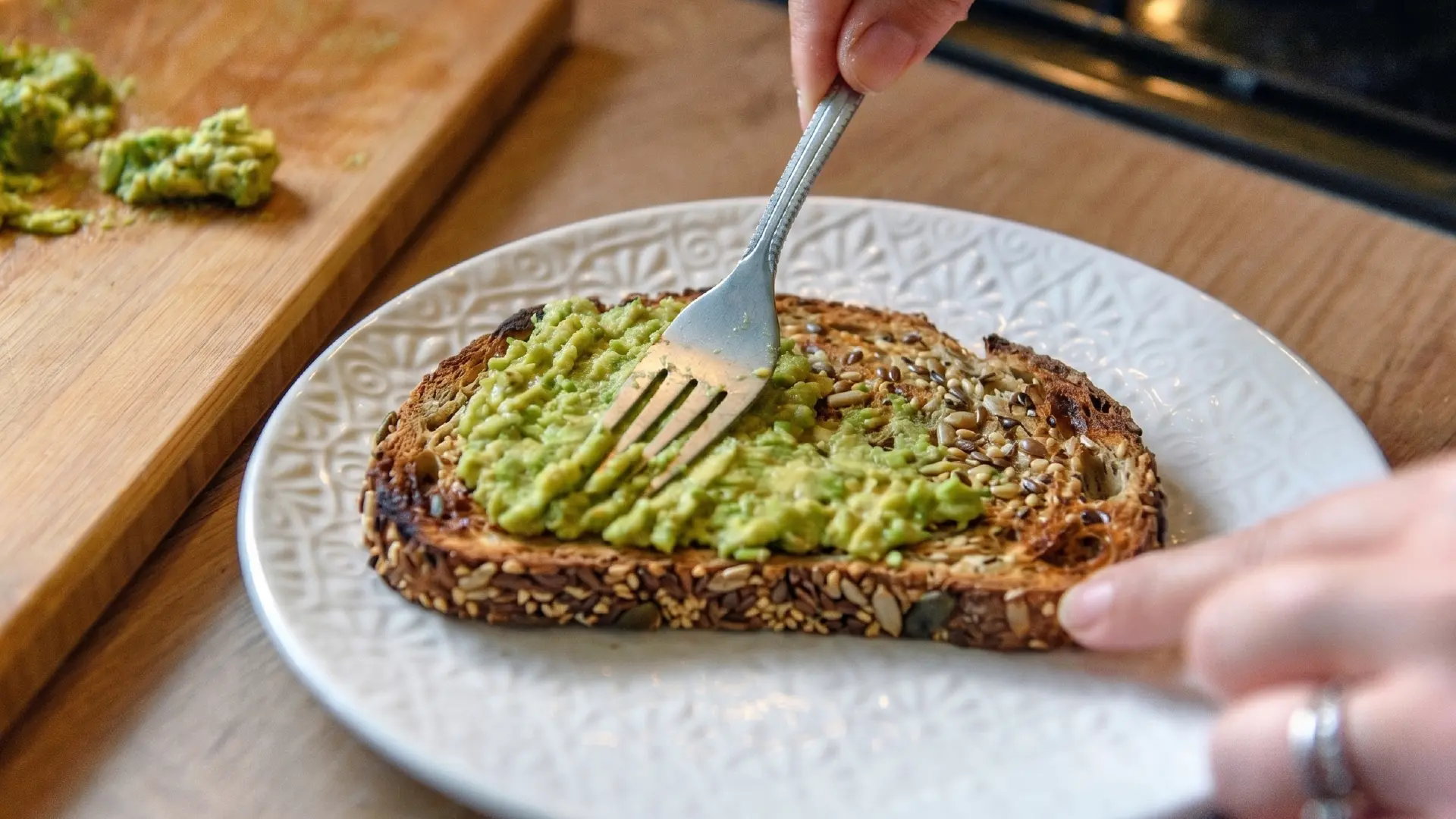 A person uses a metal fork to spread mashed avocado onto a slice of toasted seeded sourdough bread on a white plate.