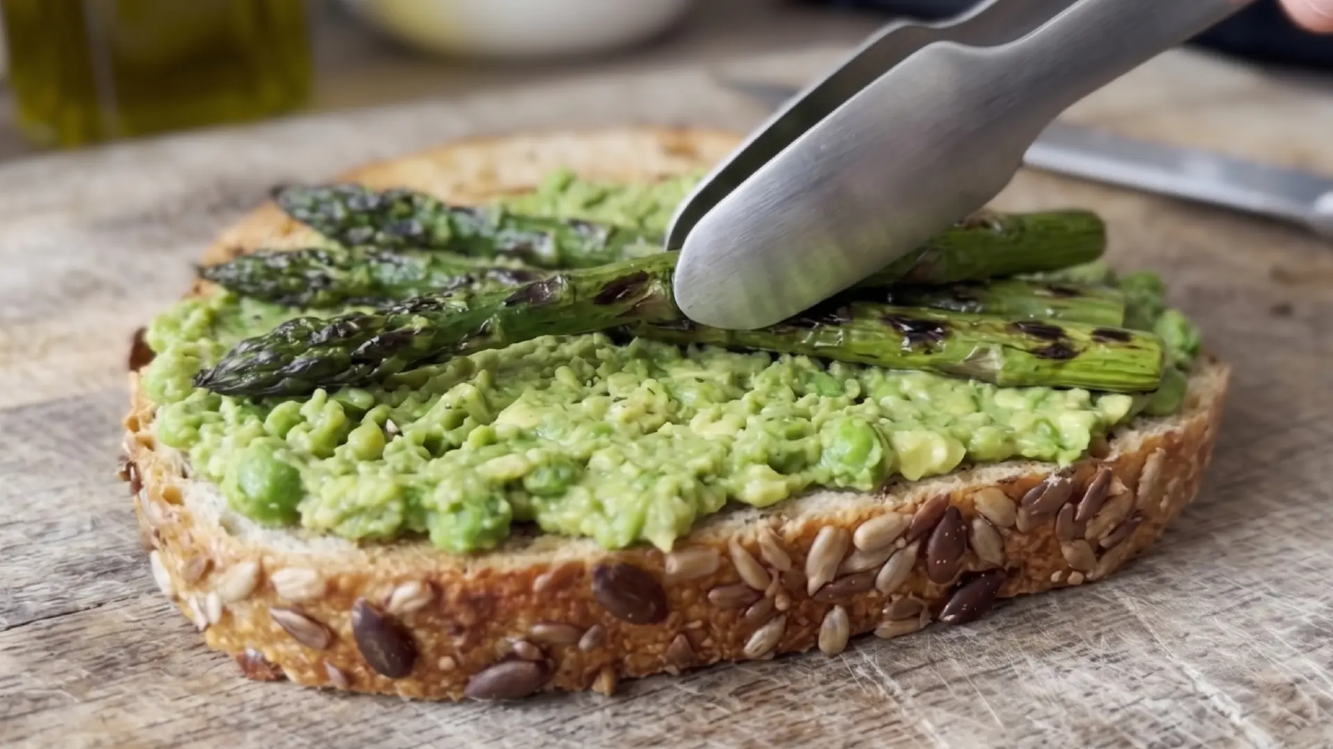 A person uses a metal fork to spread mashed avocado onto a slice of toasted seeded sourdough bread on a white plate.