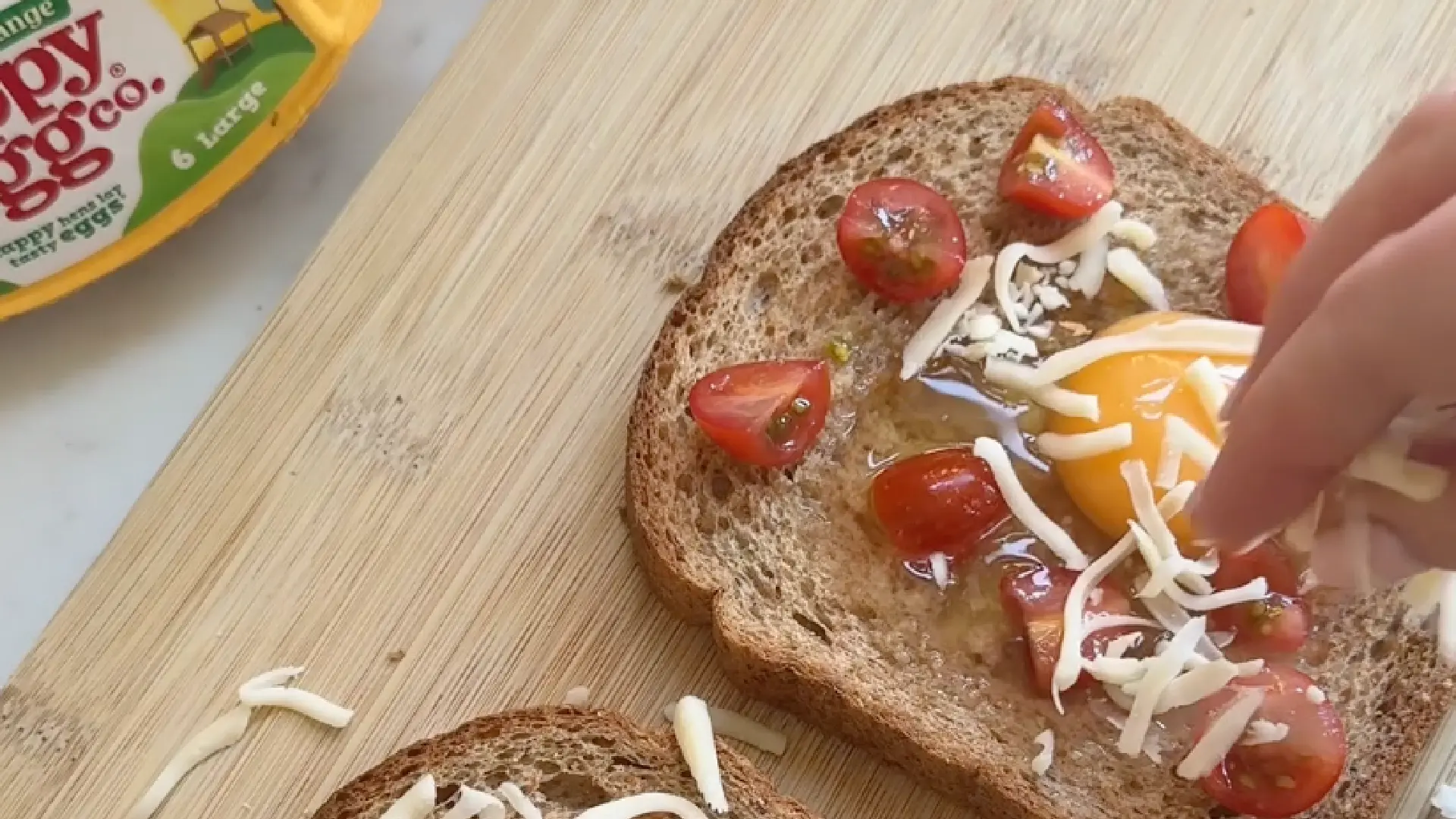Sprinkling shredded cheese and chopped cherry tomatoes over a raw egg sitting in a slice of wholemeal bread.