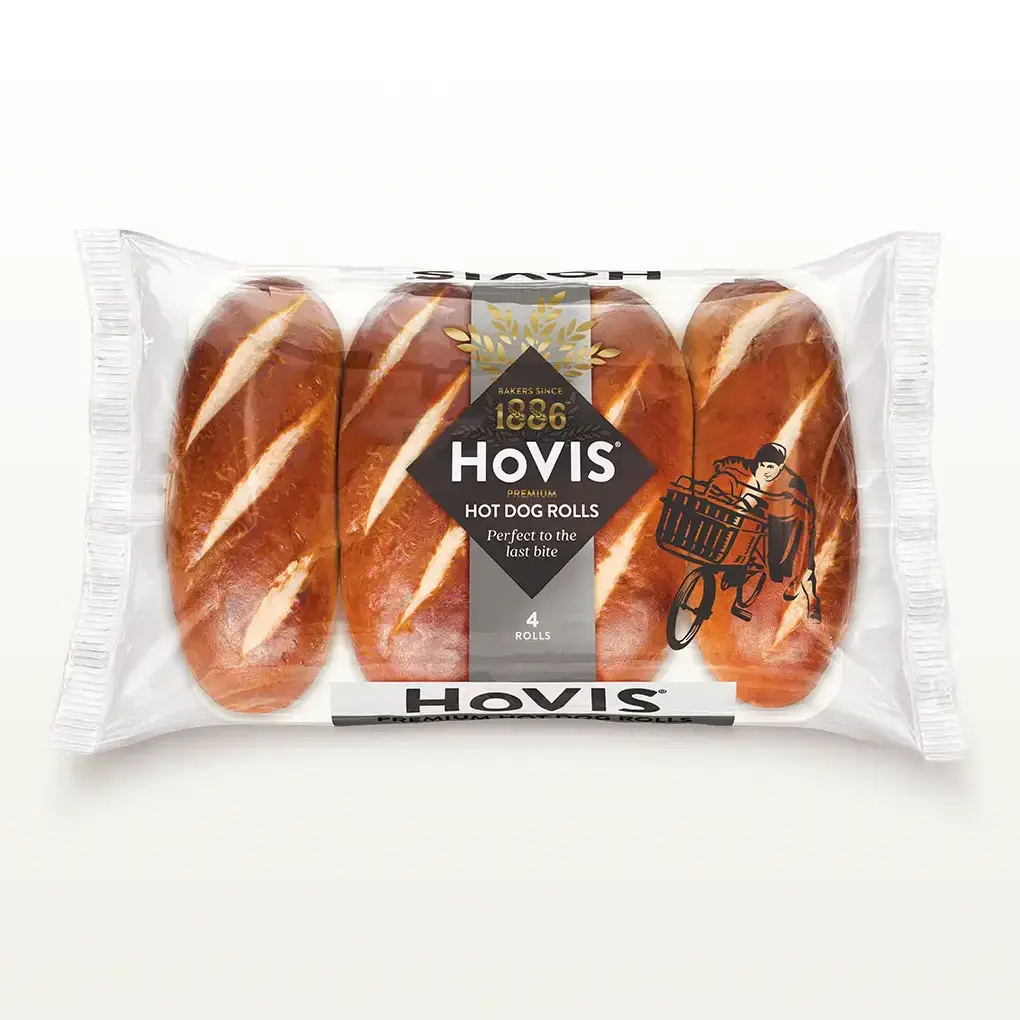 Four Hovis Hot Dog Rolls in a transparent rectangular packaging featuring the Boy on the Bike, a grey stripe, golden wheat, and the Hovis logo on top.
