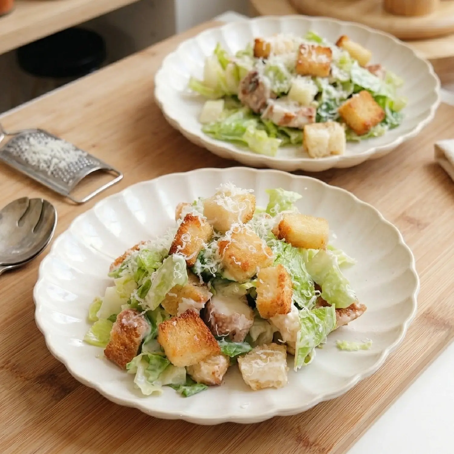 Two plates of Caesar salad with large golden croutons and shaved parmesan on a wooden board in a bright kitchen.