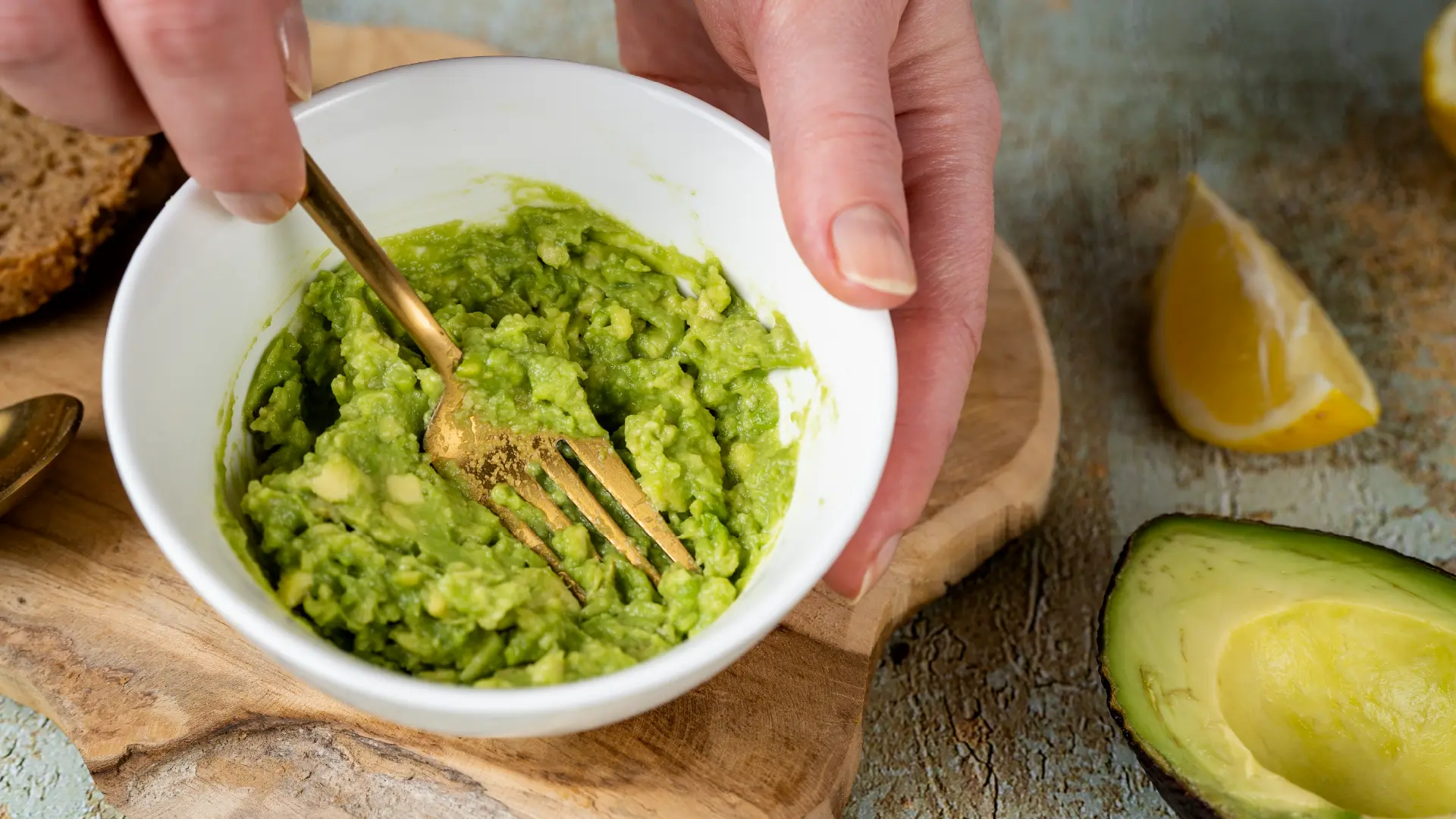 A close-up of a white bowl filled with mashed avocado being mixed by a golden fork.