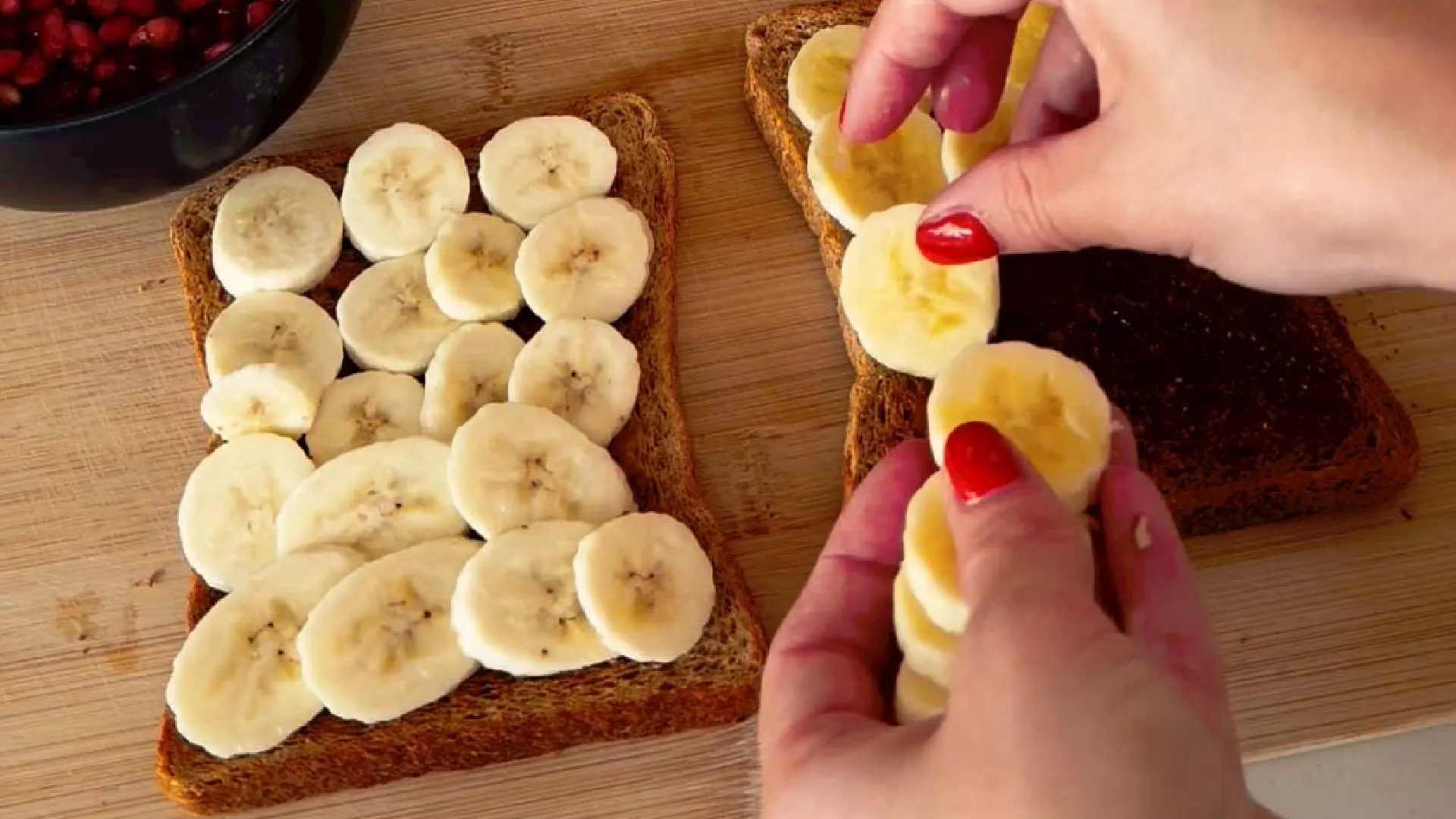 Hands layering sliced fresh bananas onto two pieces of toasted wholemeal bread on a wooden cutting board.