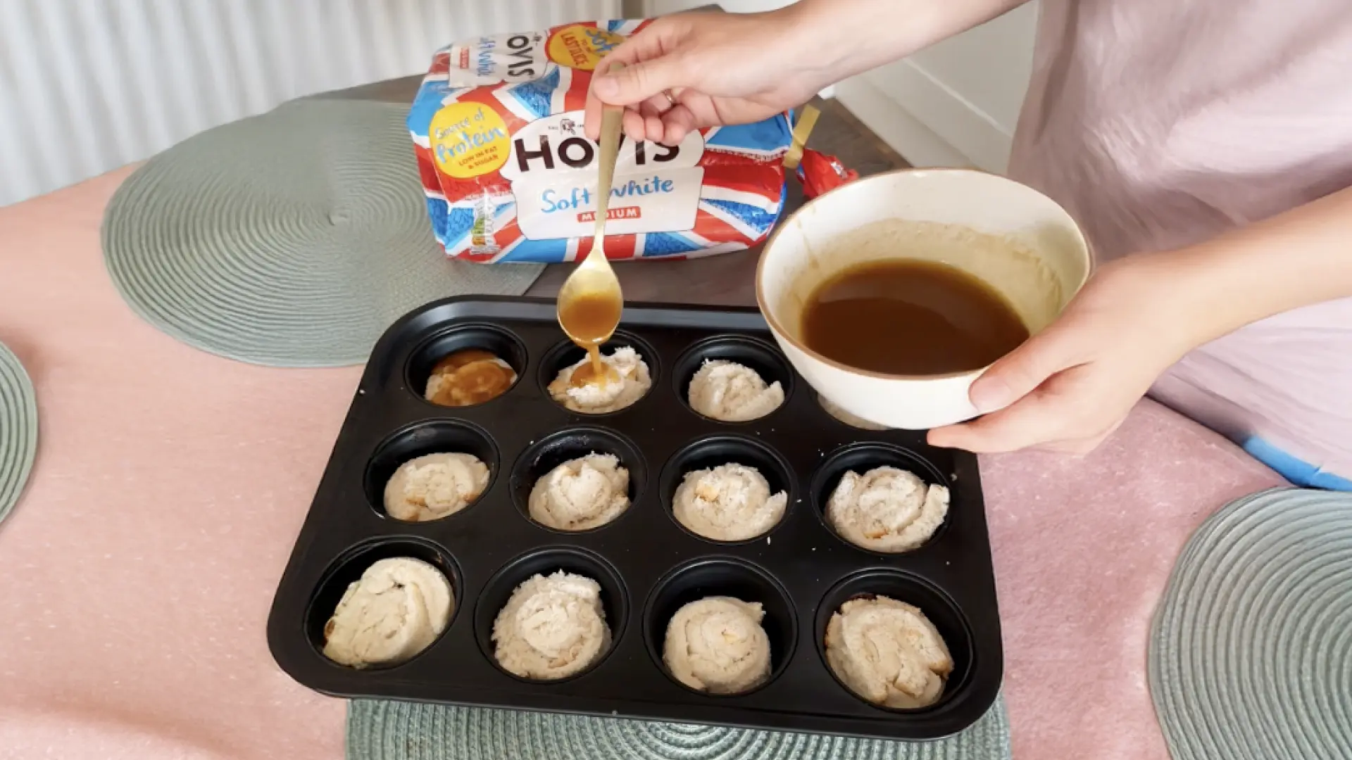 A person spoons a brown liquid over dough spirals in a muffin tin, with a Hovis bread loaf in the background.