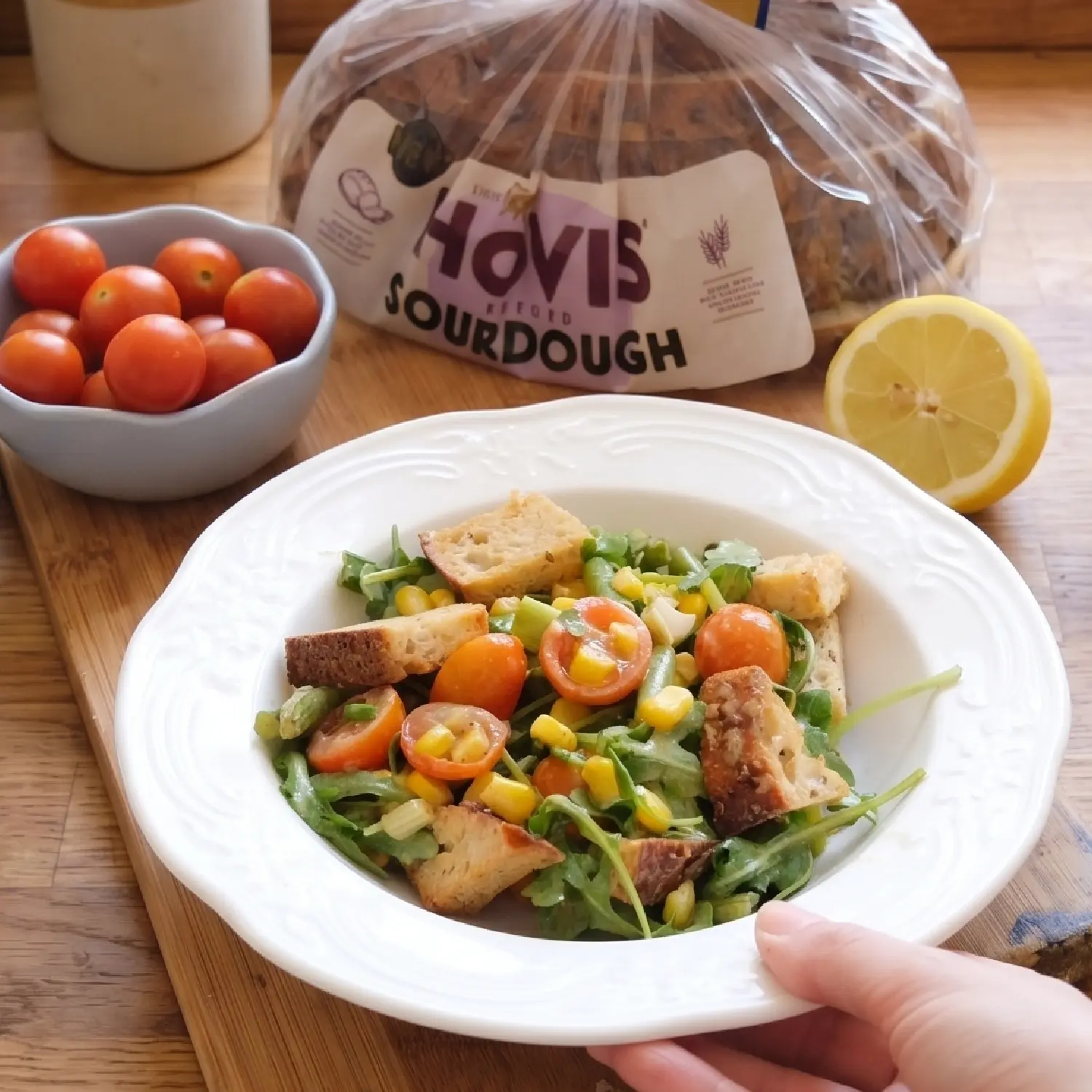 A shallow white bowl filled with a colourful salad of greens, tomatoes, corn and croutons sits on a wooden counter.