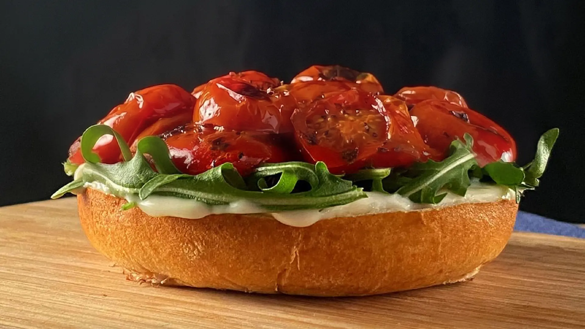 A toasted bun on a wooden board topped with white sauce, fresh green rockets leaves and juicy roasted cherry tomatoes.
