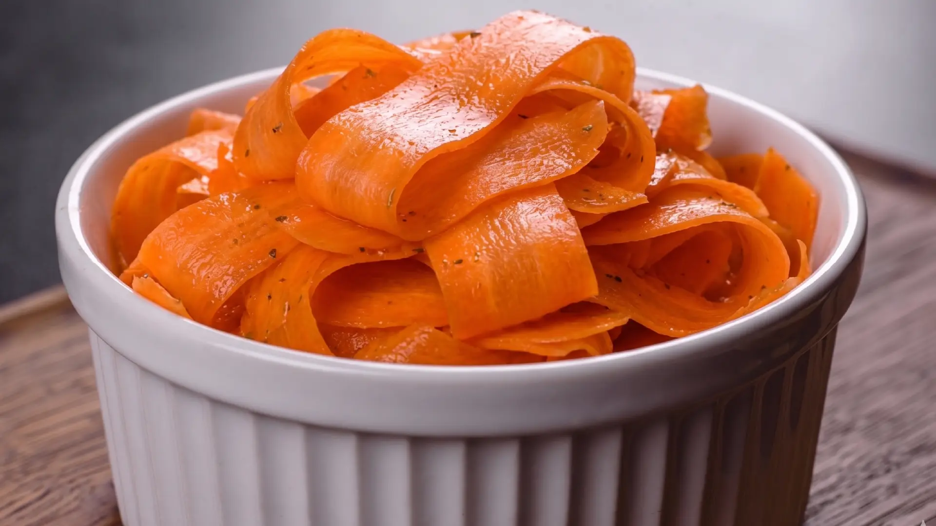 A white ramekin filled with thinly shredded, vibrant orange carrots sits on a rustic wooden surface.