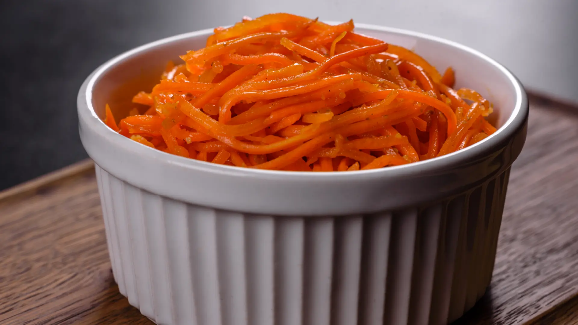 A white ramekin filled with thinly shredded, vibrant orange carrots sits on a rustic wooden surface.