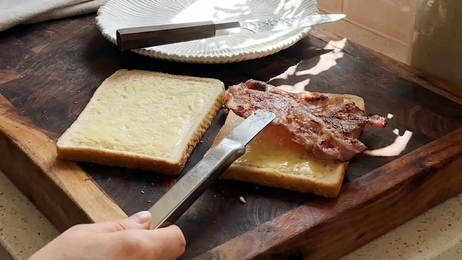 Tongs place crispy, cooked bacon onto a buttered slice of white bread to assemble a sandwich on a dark wooden surface.