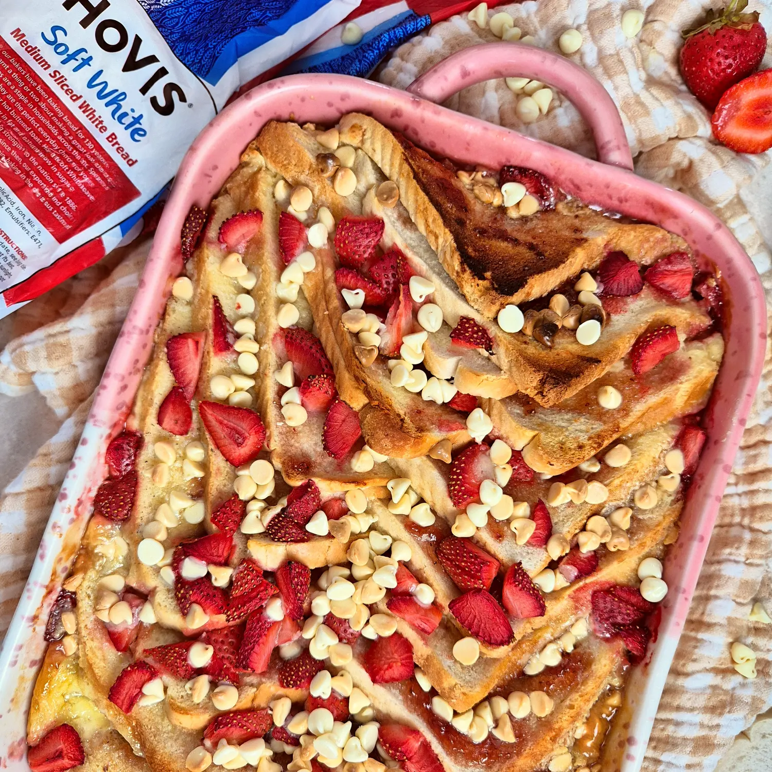 Bread and butter pudding baked in a pink dish, topped with fresh sliced strawberries and white chocolate chips.