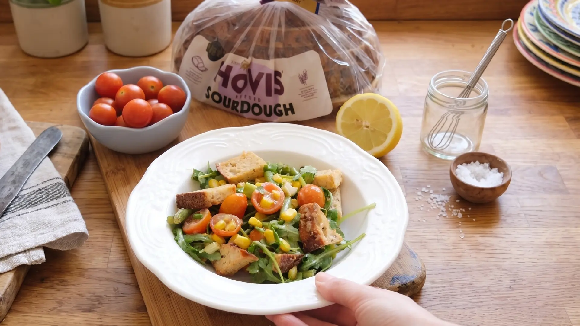 A person holds a white plate of salad on a wooden counter near a loaf of sourdough bread, tomatoes and a cut lemon.