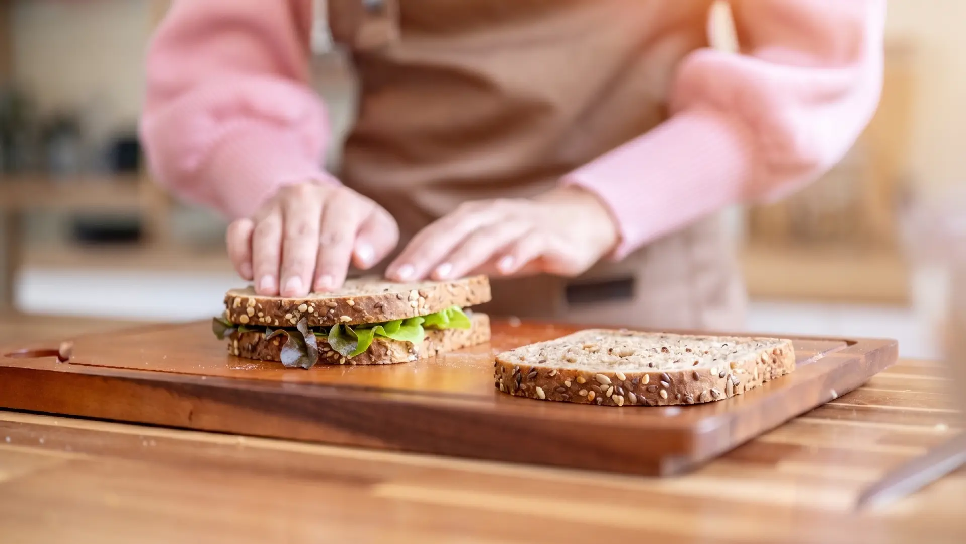 A person gently pressing down on the top slice of a freshly assembled seeded sandwich on a wooden cutting board.