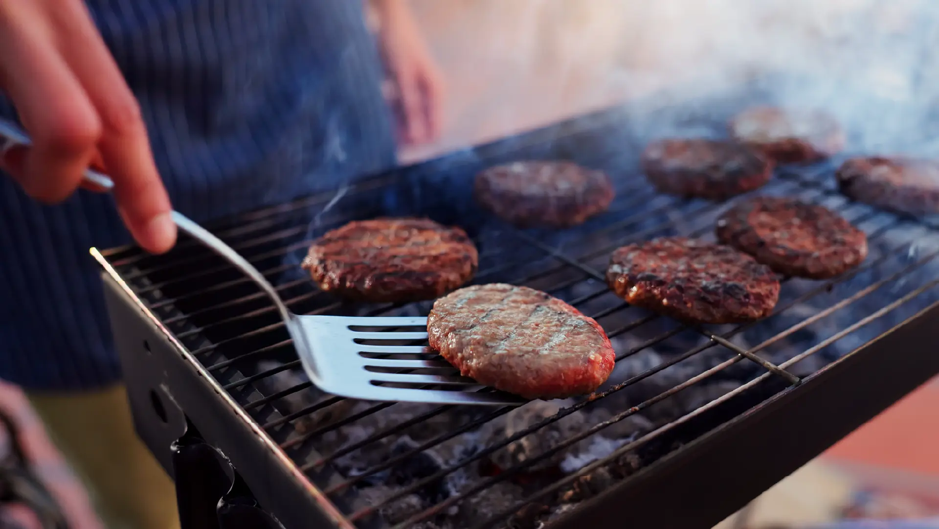 A person uses a metal spatula to flip burger patties cooking over a smoky outdoor grill.