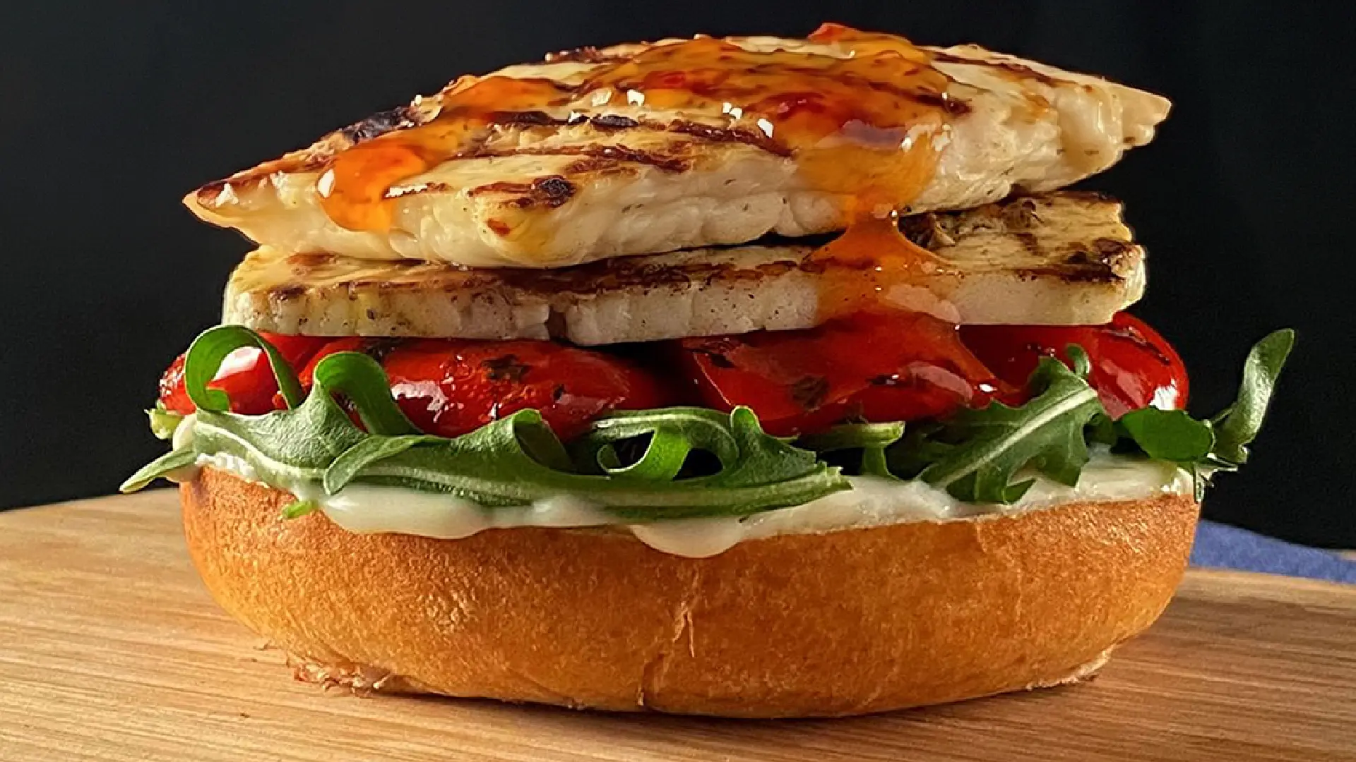 A halloumi burger on a wooden board with layers of white sauce, rocket, roasted tomatoes, grilled cheese and sweet chilli sauce.