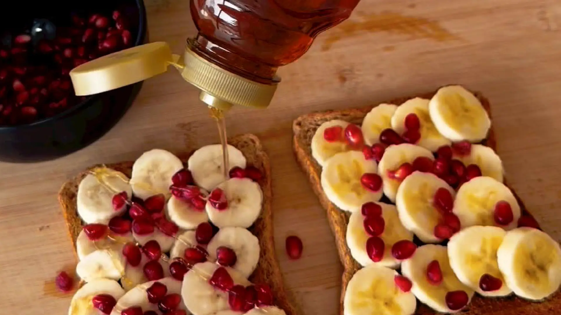 Golden honey drizzling from a squeeze bottle onto banana and pomegranate topped wholemeal toast.