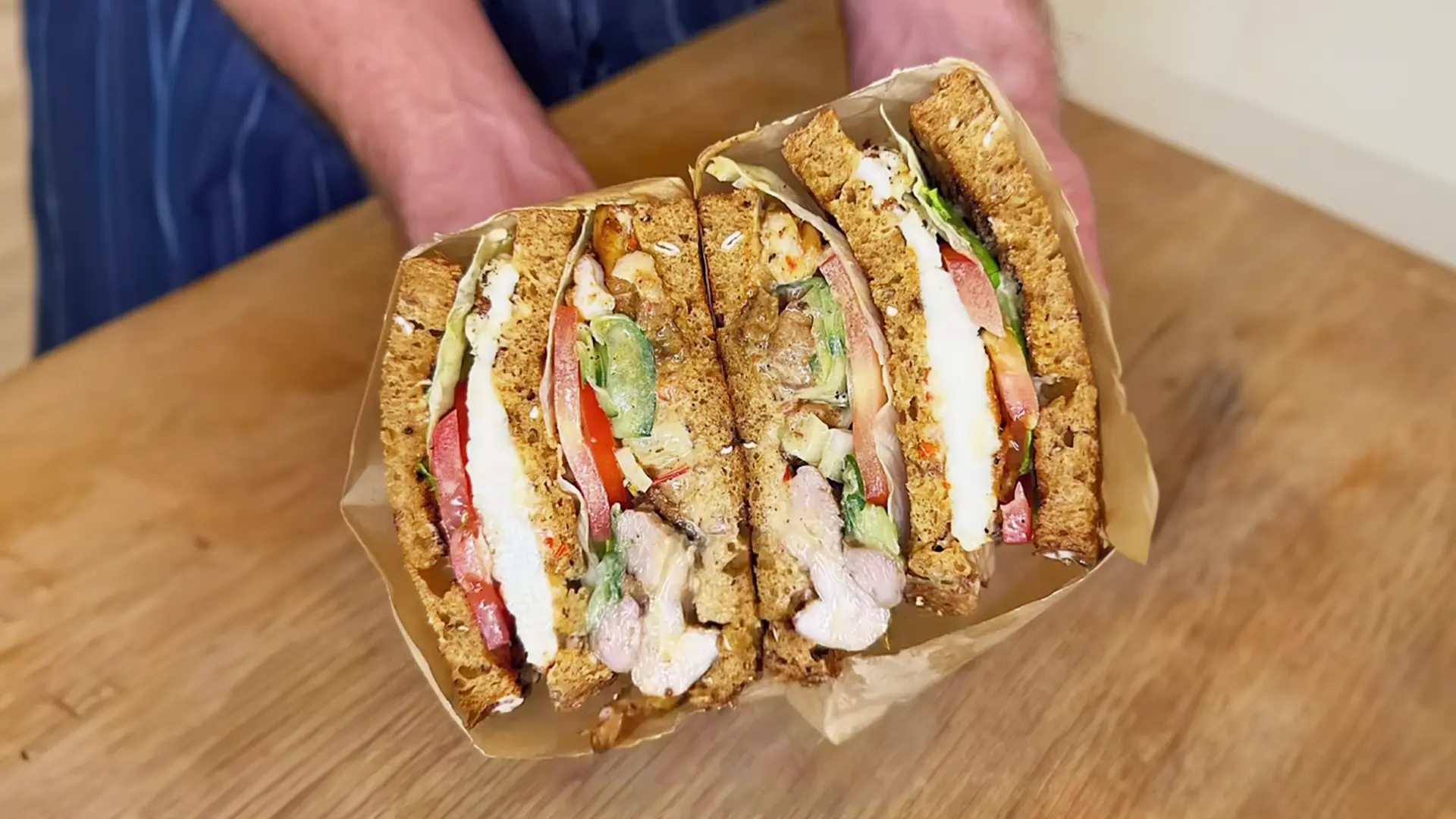 Someone holding two halves of a sandwich made with Hovis Granary Half Cob wrapped in paper. The sandwich is made of brown bread, filled with tomato slices, lettuce, juicy grilled chicken, and thin cucumber slices.