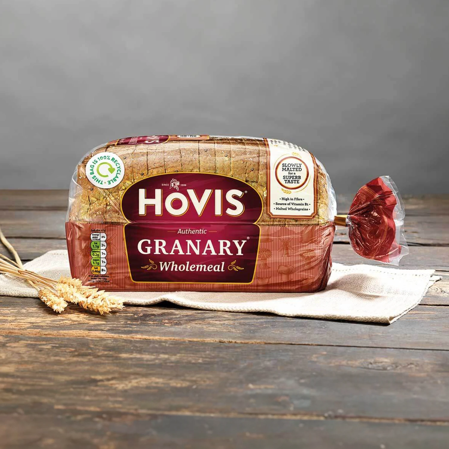 Hovis Granary 400g loaf on beige napkins against a light grey background, resting on a rustic wooden table with natural light, with a couple of dried wheat stalks on the left.