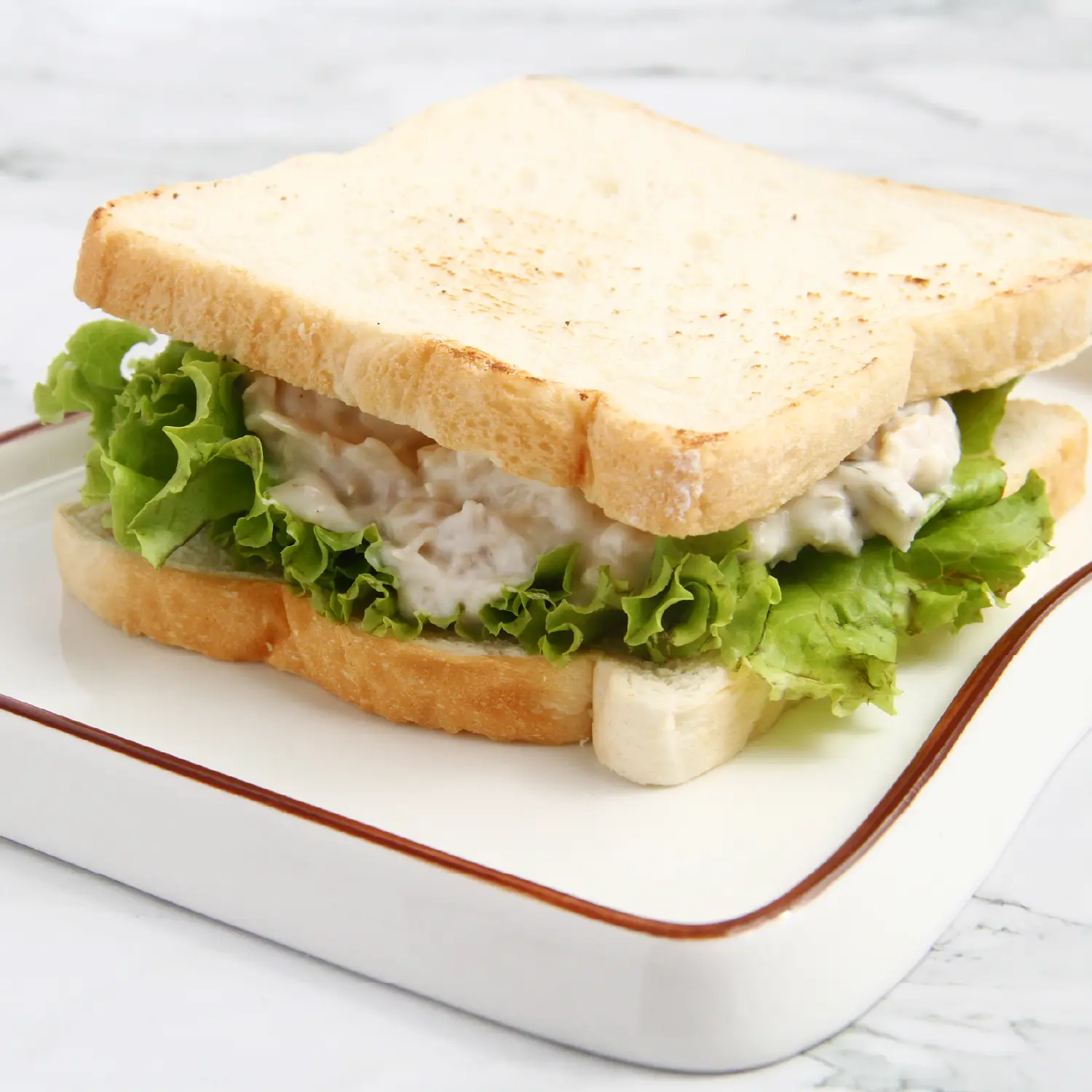A chicken salad sandwich with green lettuce on white bread, served on a square white plate with a brown rim.