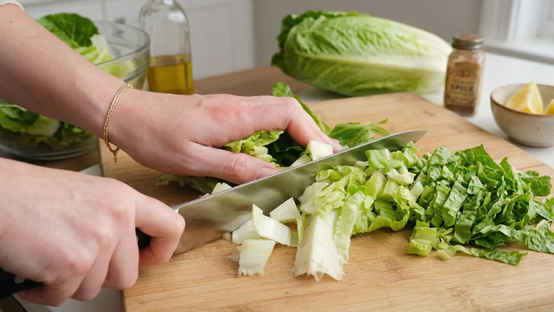Close-up of a person's hands using a chef's knife to chop fresh romaine lettuce on a wooden cutting board in a bright kitchen.