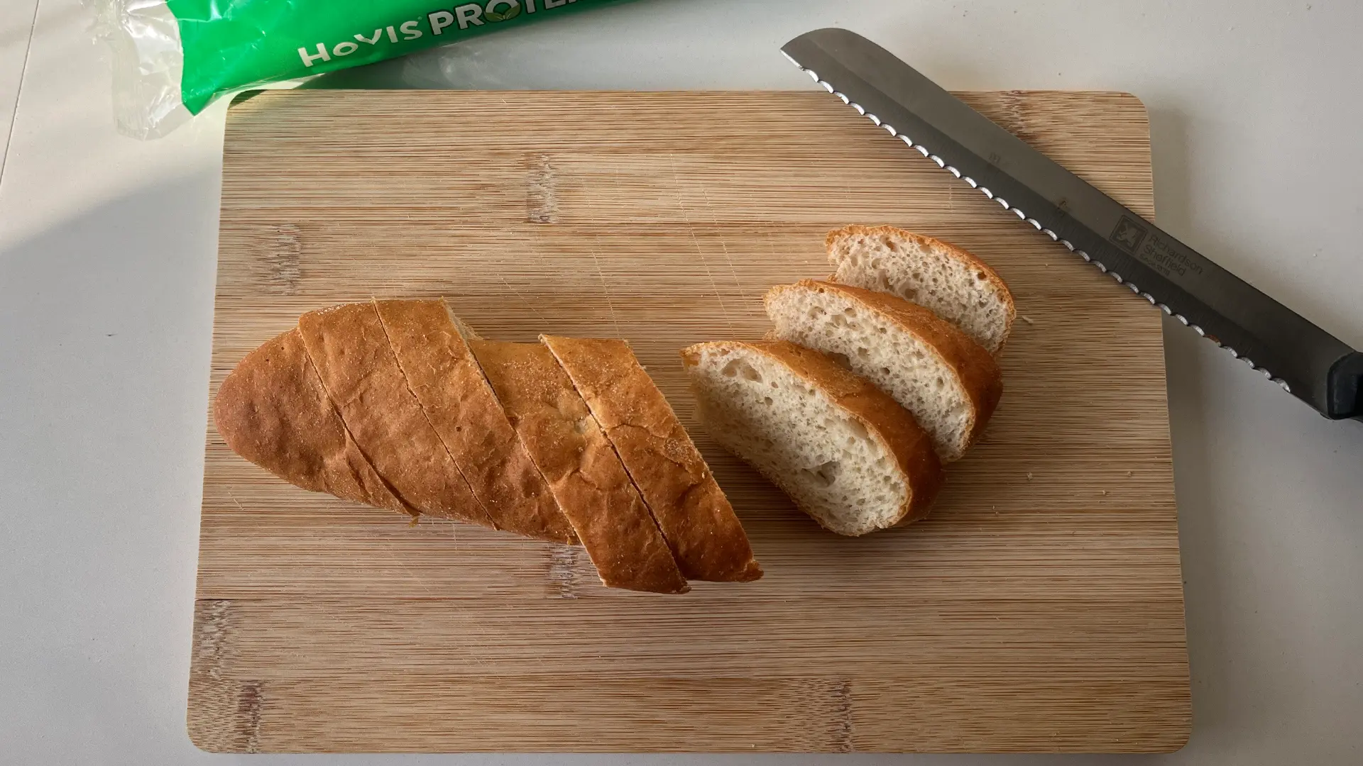 A top-down shot of a wooden chopping board with a serrated knife resting on the upper right side. On the board are several slanted slices of a golden-brown sub roll. The slices are arranged in two rows, with the remaining loaf partially visible. A green bag of Hovis bread is blurred in the top background.