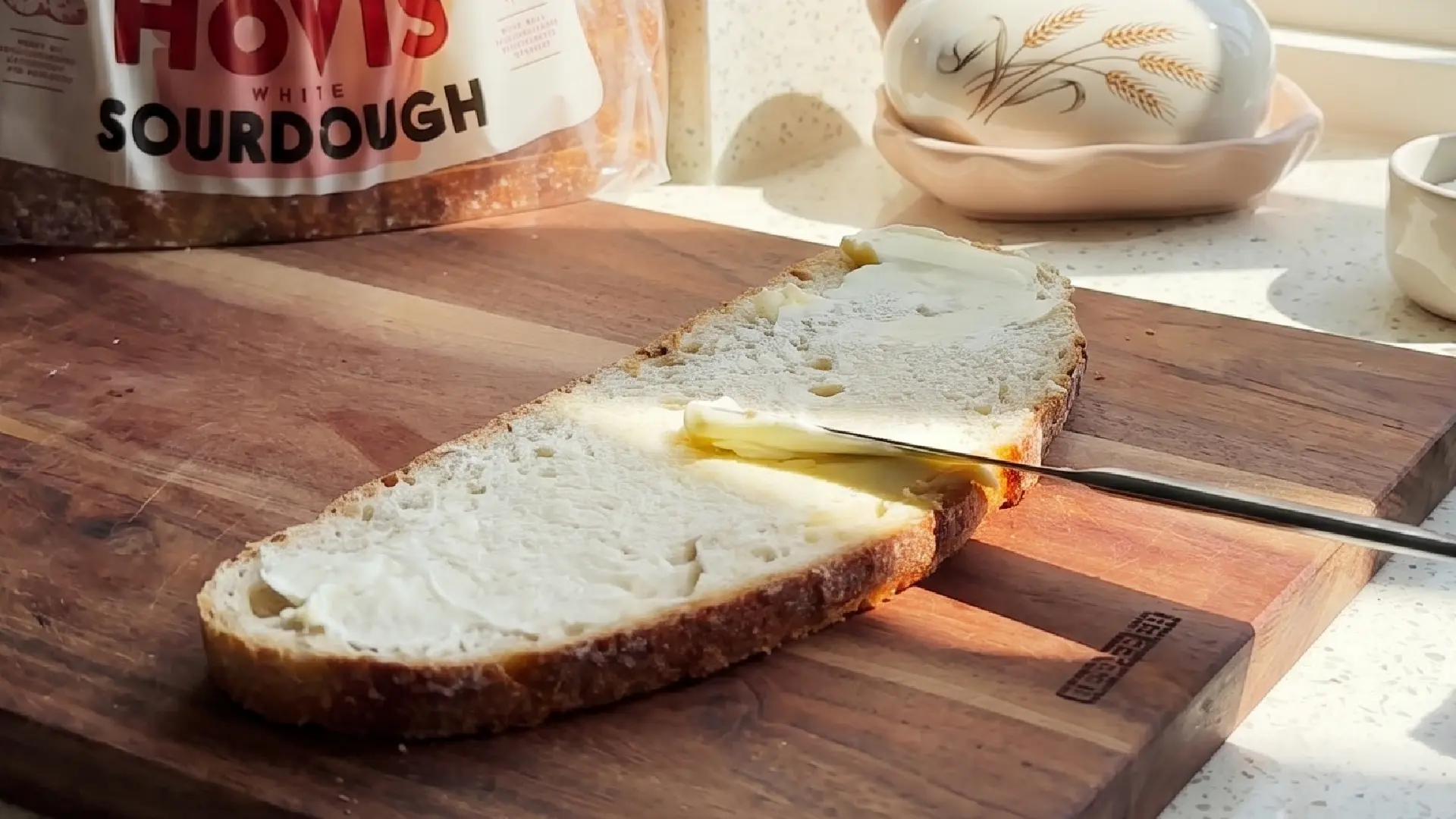 A slice of sourdough bread is spread with butter on a wooden cutting board next to a Hovis bread bag.