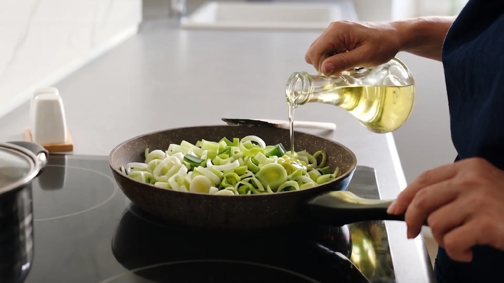 A person pours oil from a glass carafe into a skillet filled with chopped leeks on a modern glass stovetop.